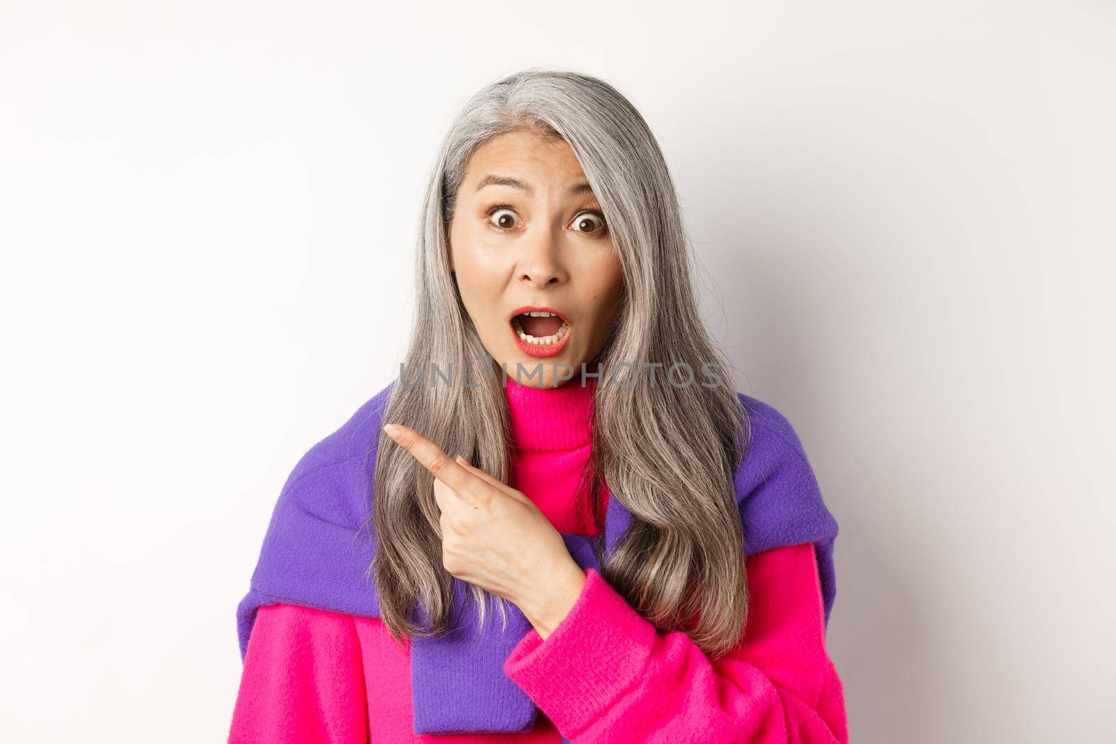 Close-up of amazed and intrigued asian senior woman pointing finger left at something cool, staring at camera impressed, standing over white background by Benzoix