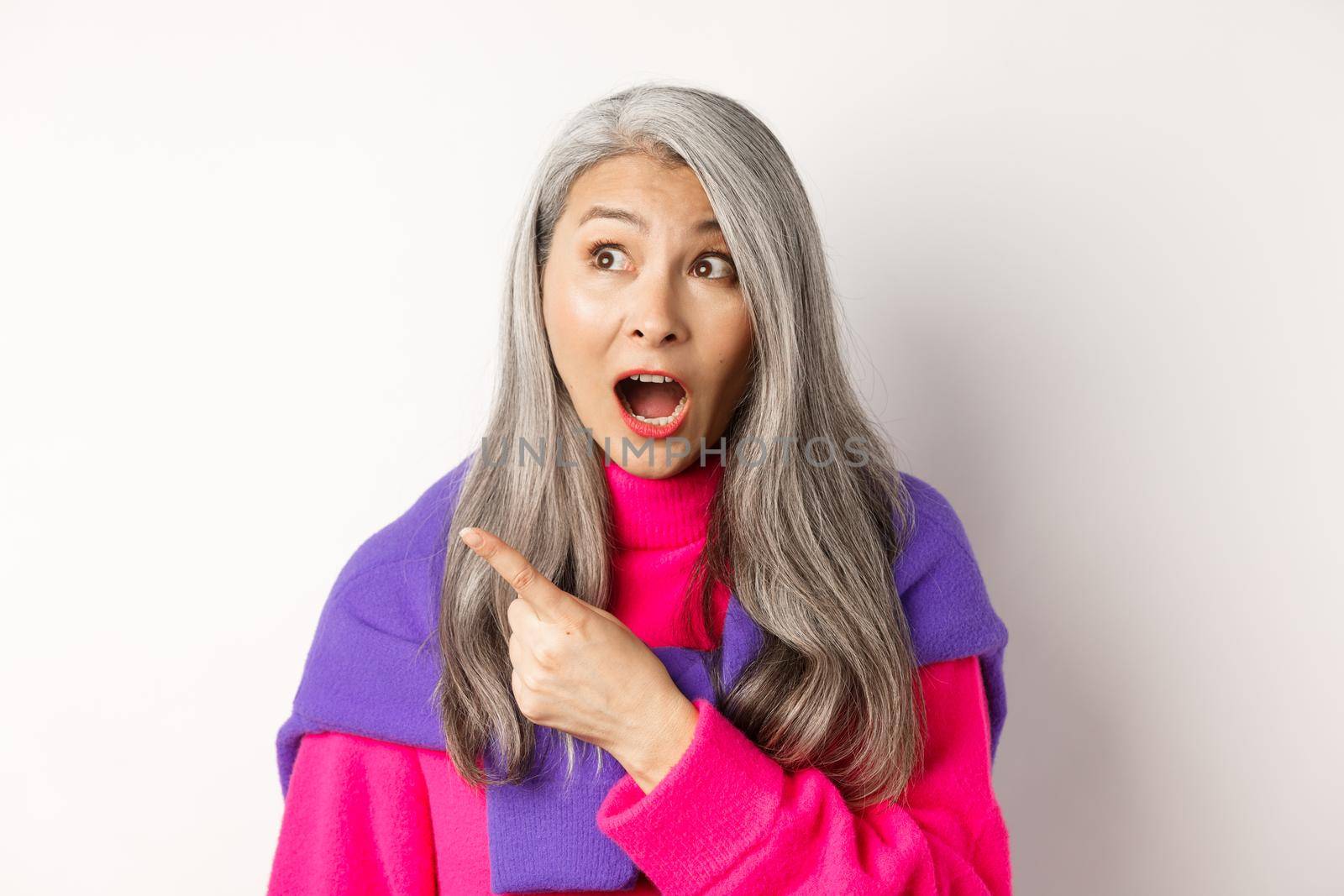 Close-up of amazed and intrigued asian senior woman pointing finger left at something cool, staring at camera promo offer, standing over white background by Benzoix