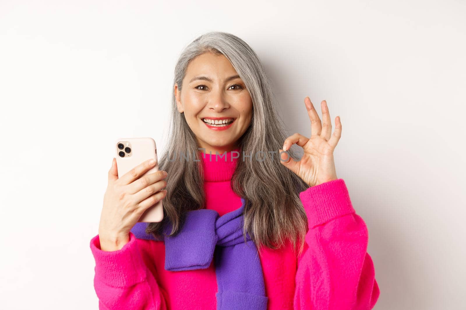 Online shopping. Close up of fashionable asian senior woman showing okay sign, using smartphone and approve something, standing over white background.