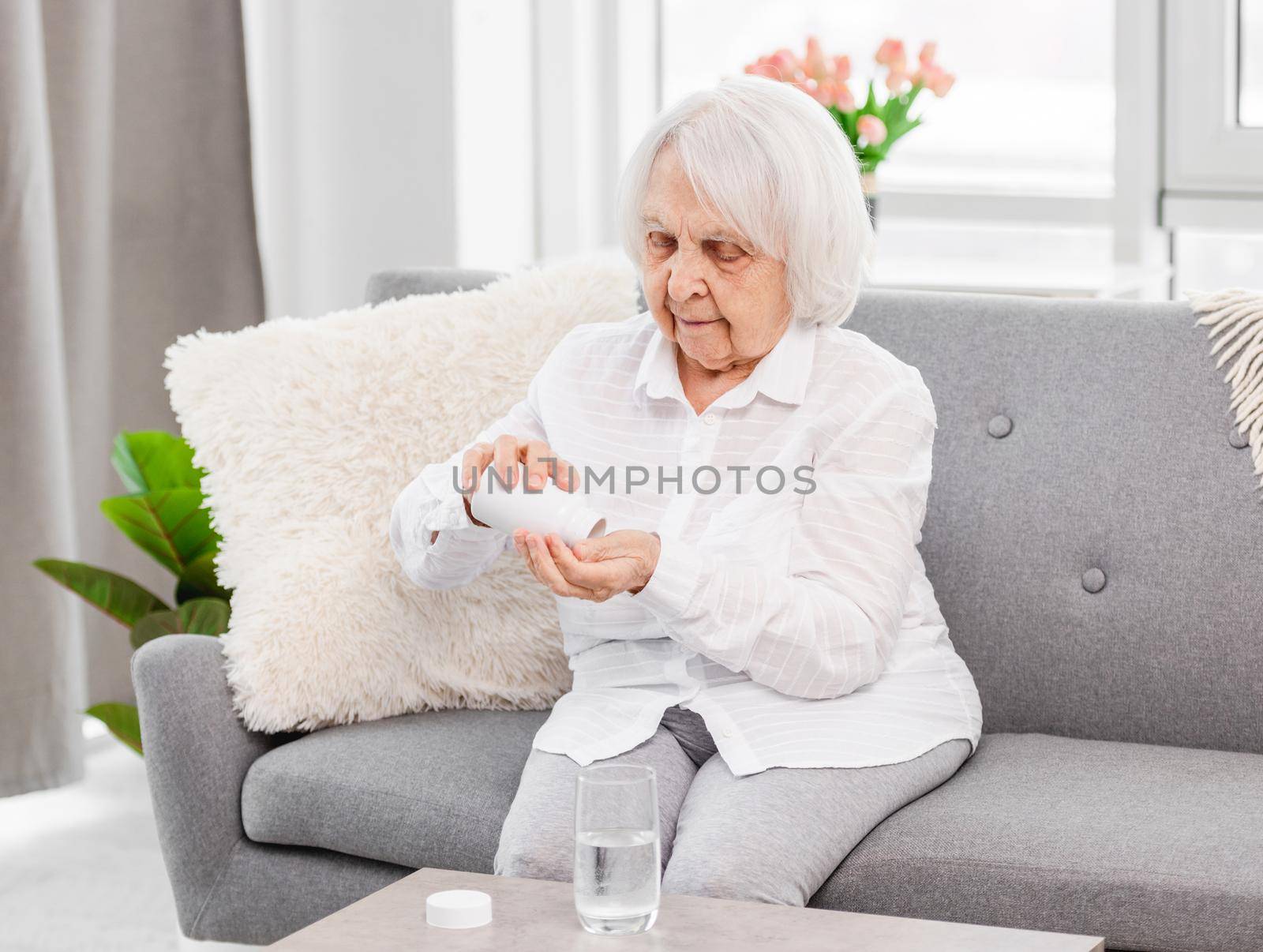 Eldery woman sitting on the sofa and taking pills at home. Pensioner with medicaments