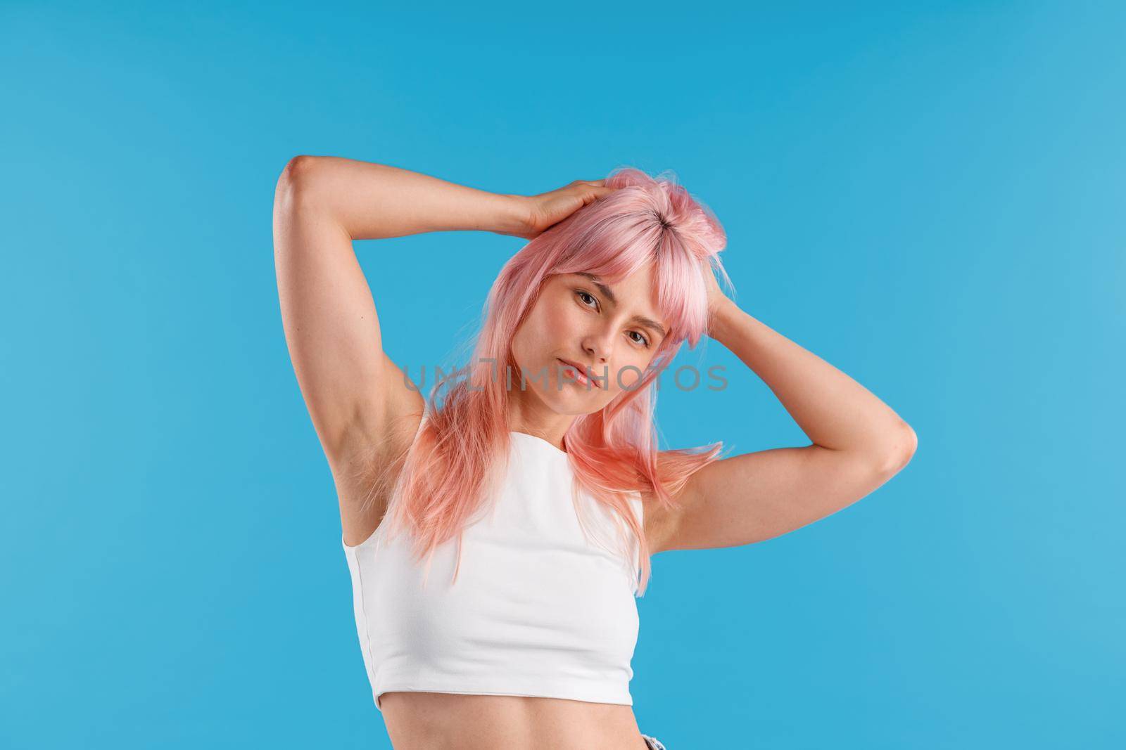 Attractive young woman in white shirt looking at camera, adjusting her pink hair while posing isolated over blue studio background. Beauty, hair care concept