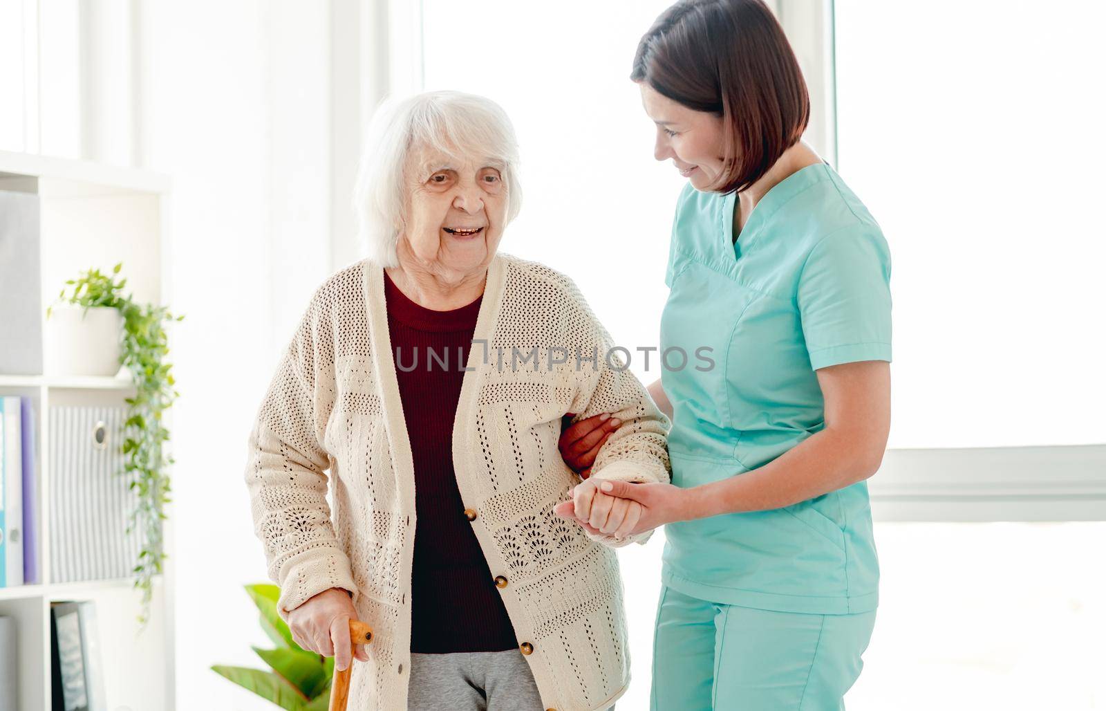 Smiling caregiver helping happy elderly woman to walk in nursing home