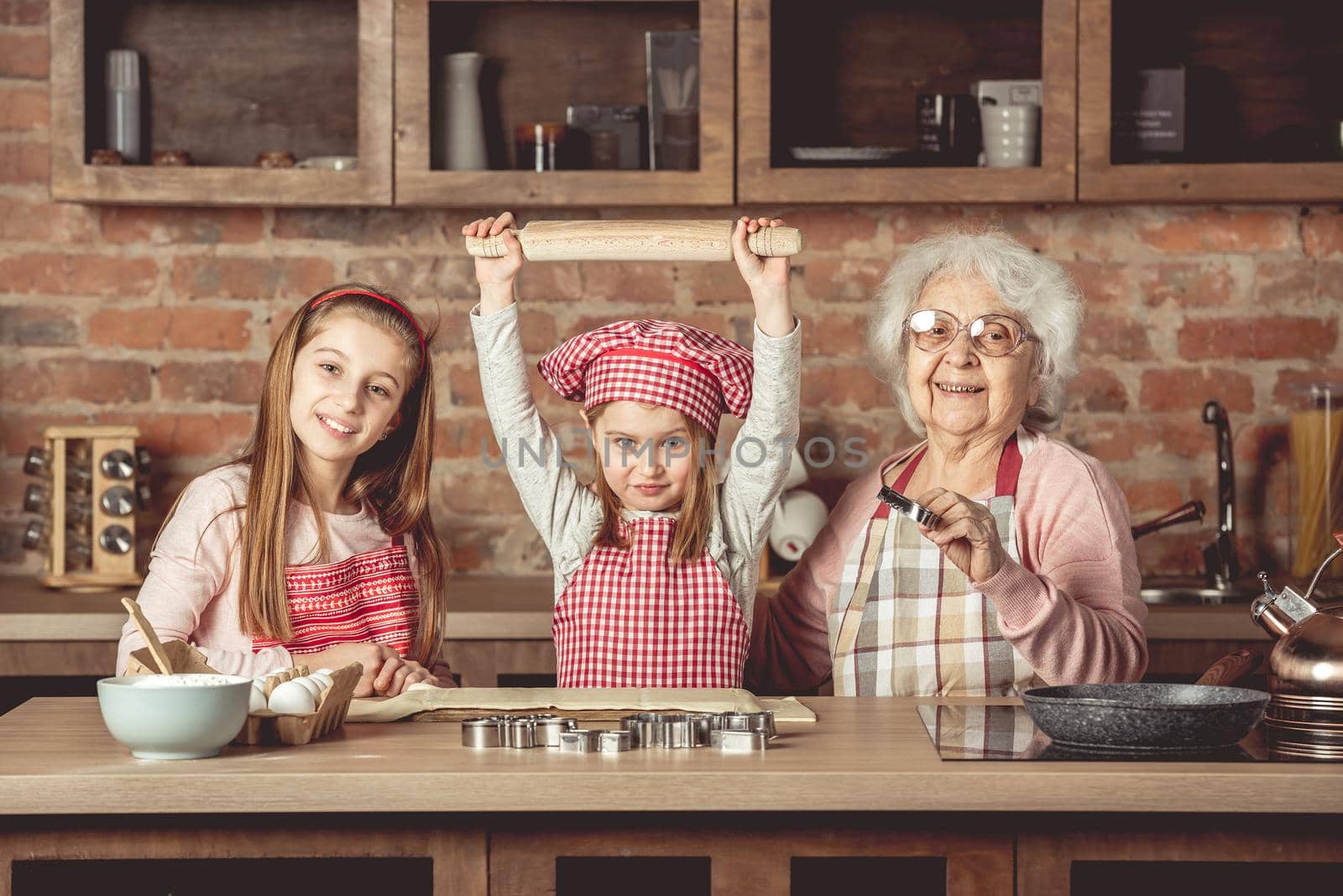 Two little grandchilds in red aprons and cooking hats ready to make homemade cookies with their eldery grandmother