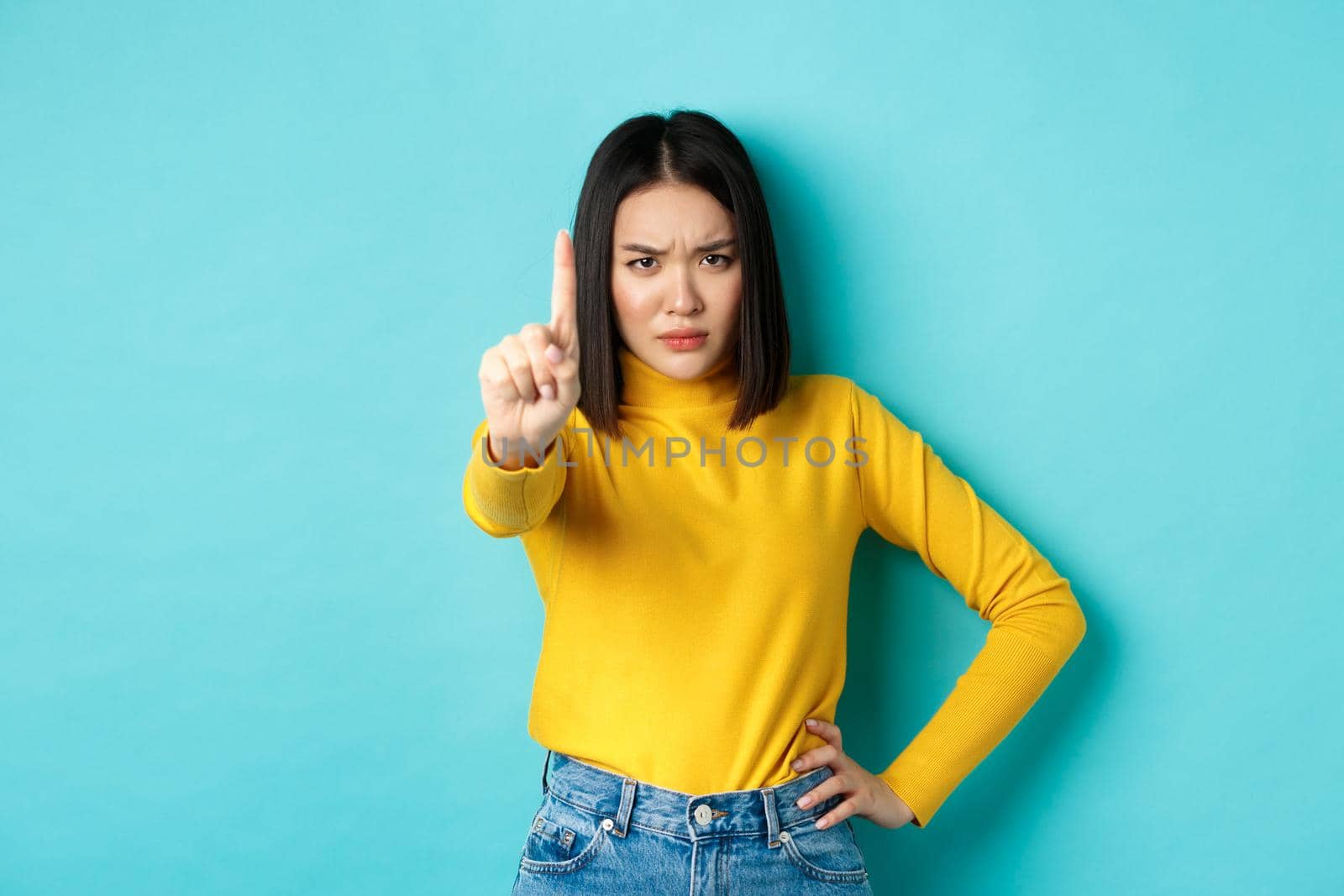 Confident and serious woman tell no, showing extended finger to stop and prohibit something bad, frowning and looking at camera self-assured, standing over blue background.