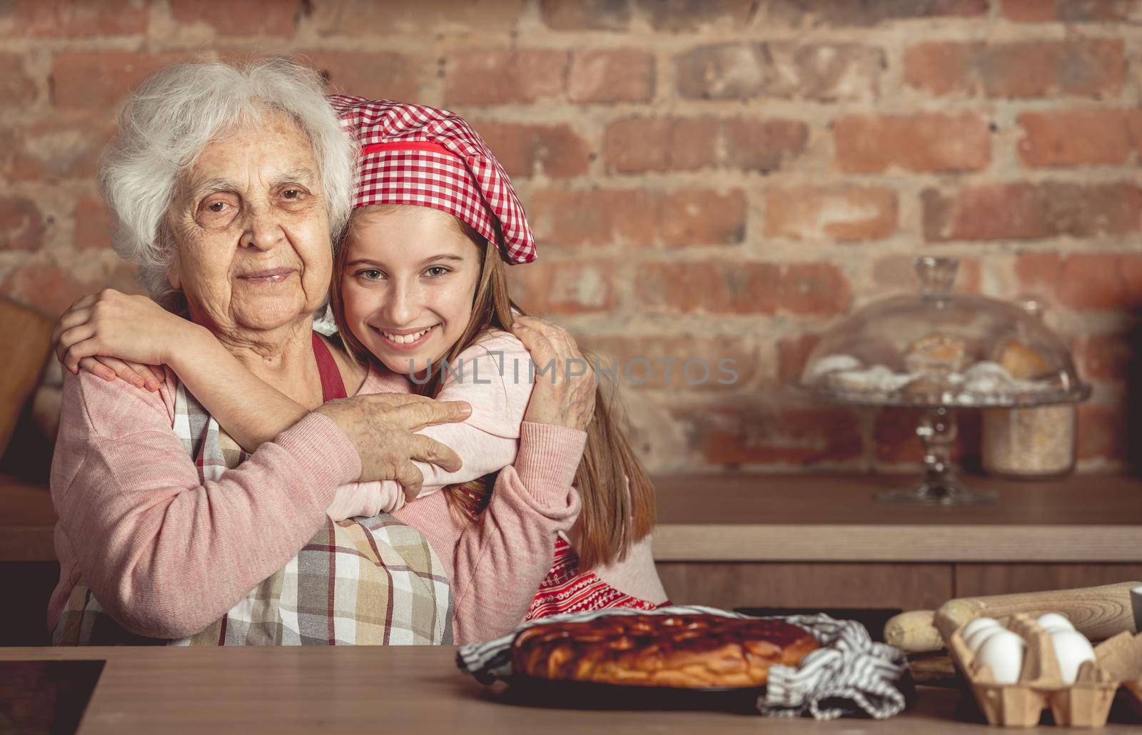 Little granddaughter hug her happy grandmother sitting at rustic kitchen at home