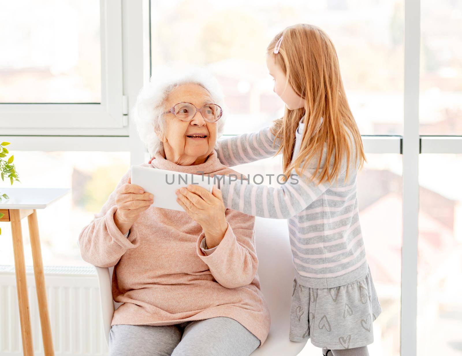 Nice girl helping to grandmother to use tablet indoors