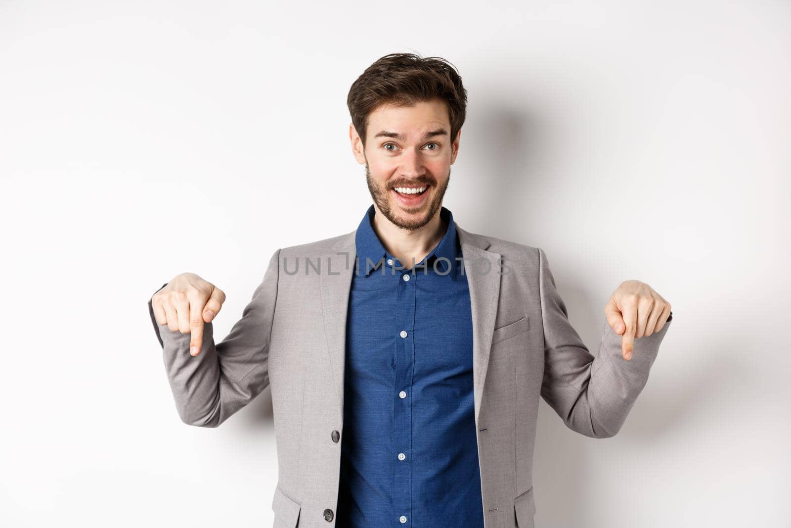 Hey check this out. Happy businessman in suit pointing fingers down and smiling, inviting to event, showing link, standing on white background by Benzoix