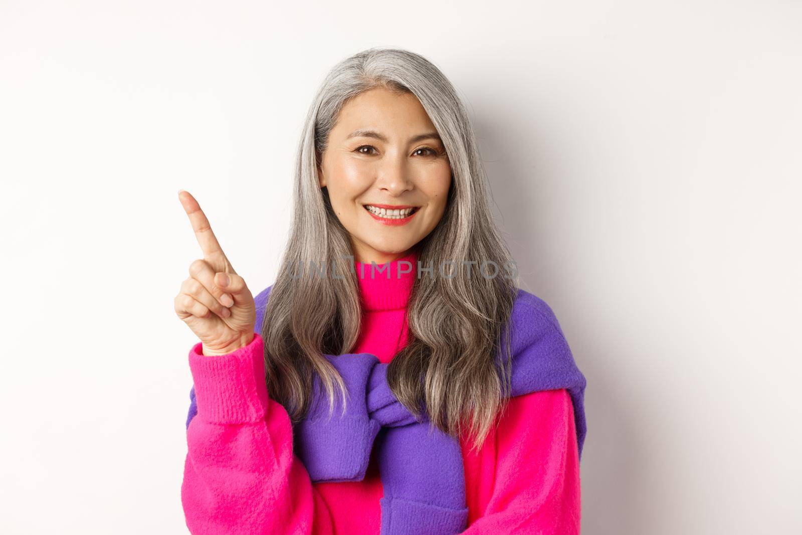 Smiling asian senior woman showing promo offer, looking happy and satisfied while pointing upper left corner, standing over white background.