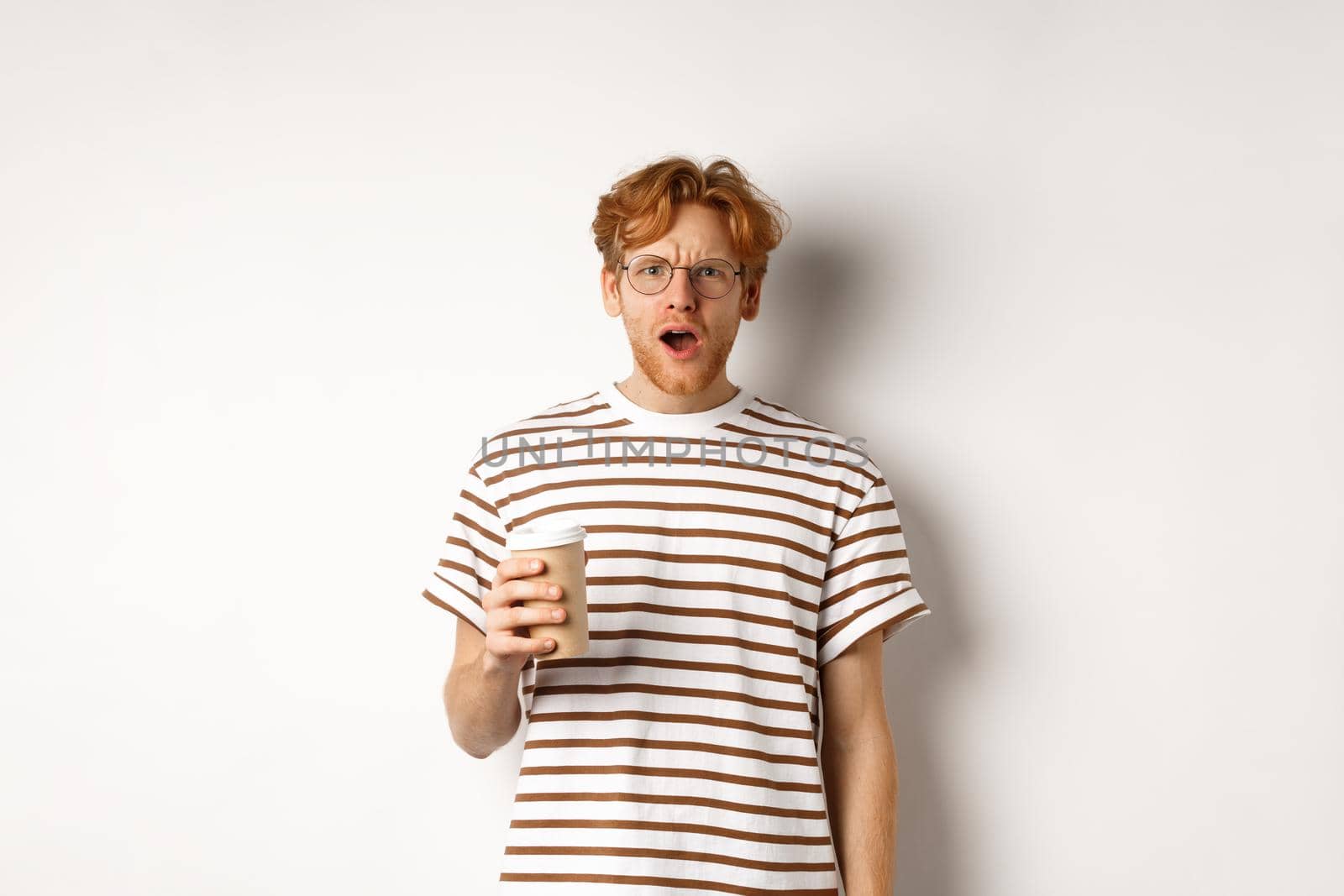Amazed redhead man in glasses holding coffee cup and staring at camera with complete disbelief, standing in striped t-shirt against white background.