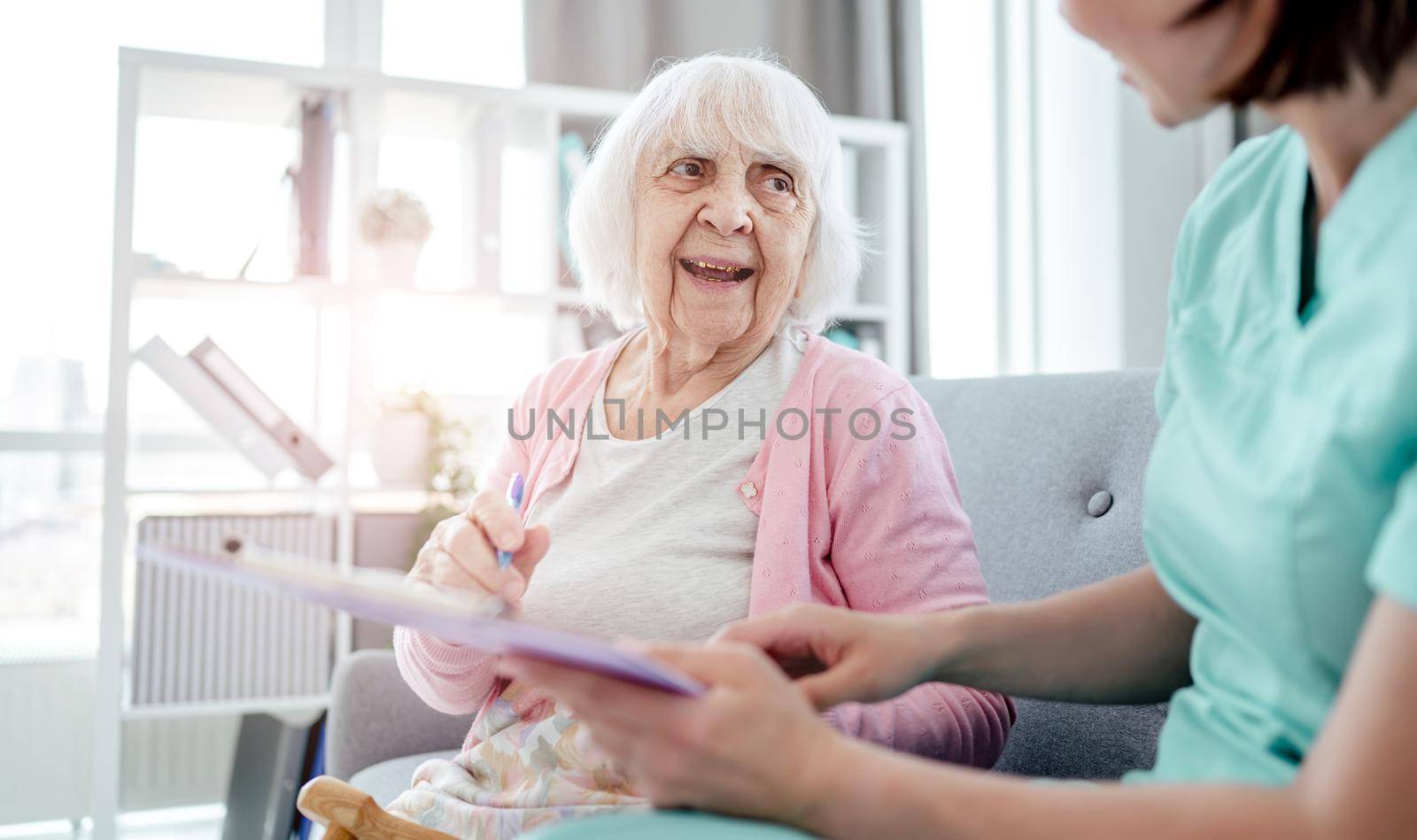 Elderly woman and nurse sign medical documents at home. Healthcare worker girl and senior female person indoors