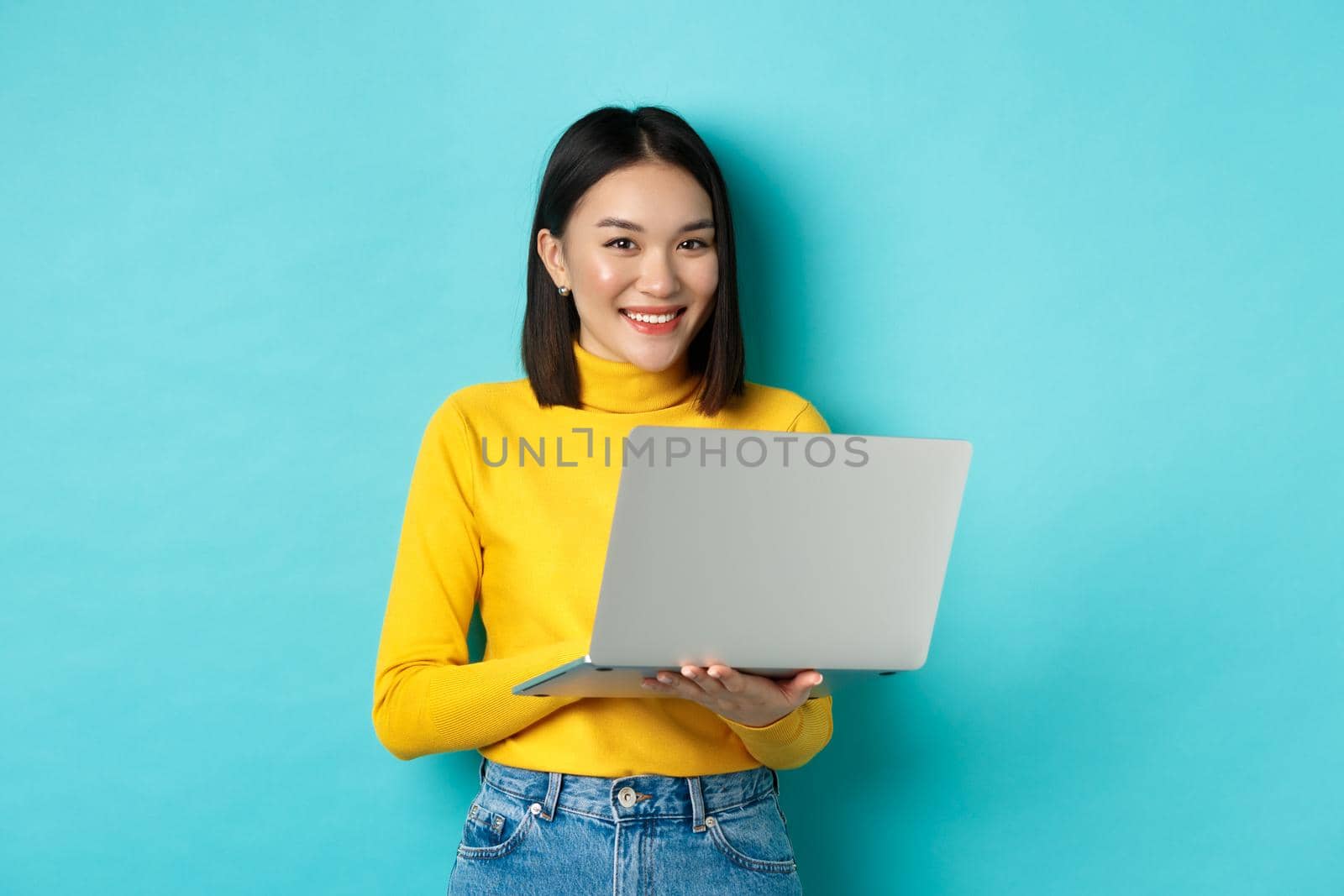 Happy asian woman in yellow pullover using laptop, shopping online or working, standing over blue background.