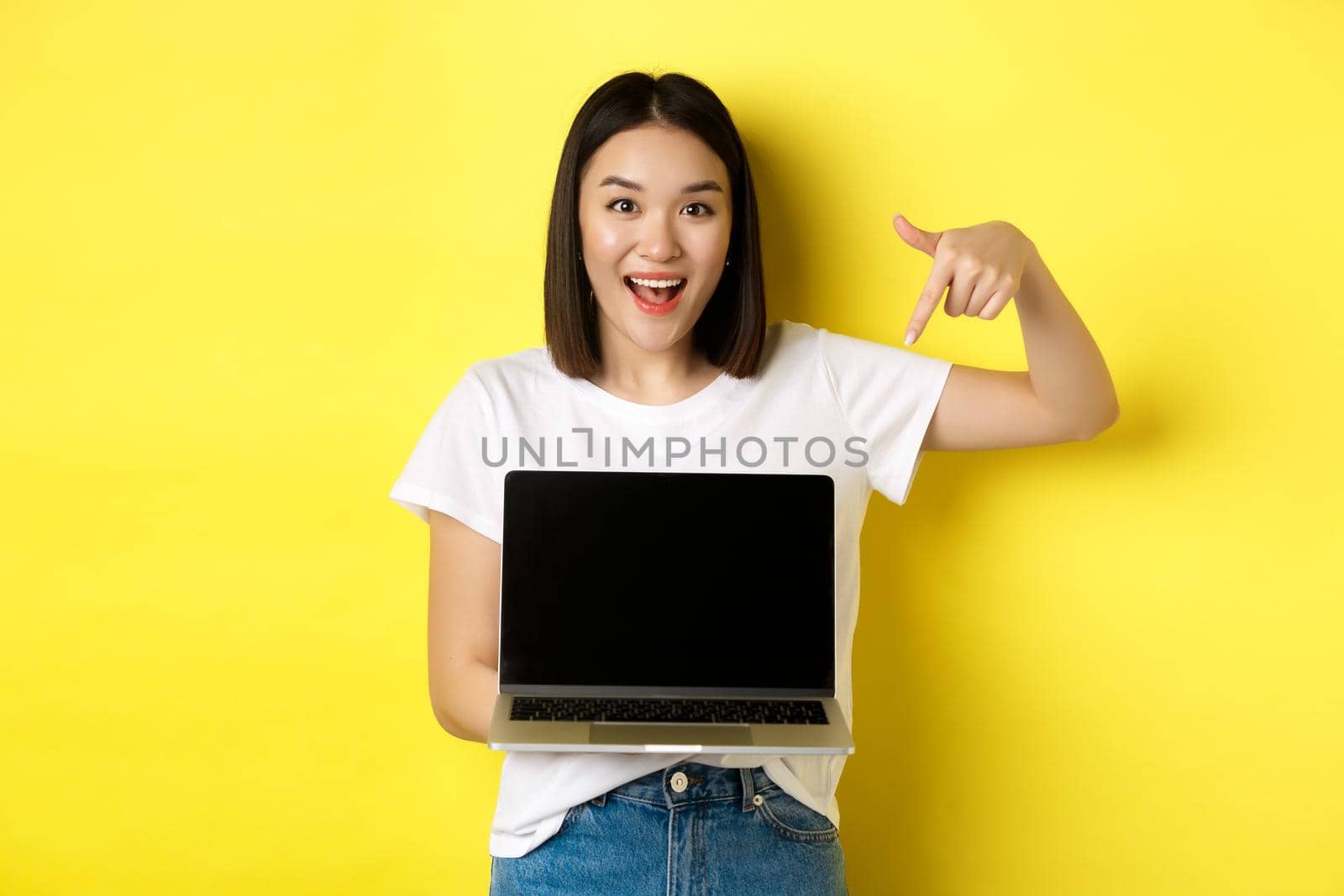 Young asian woman demonstrate online offer, showing laptop screen and smiling, standing over yellow background by Benzoix
