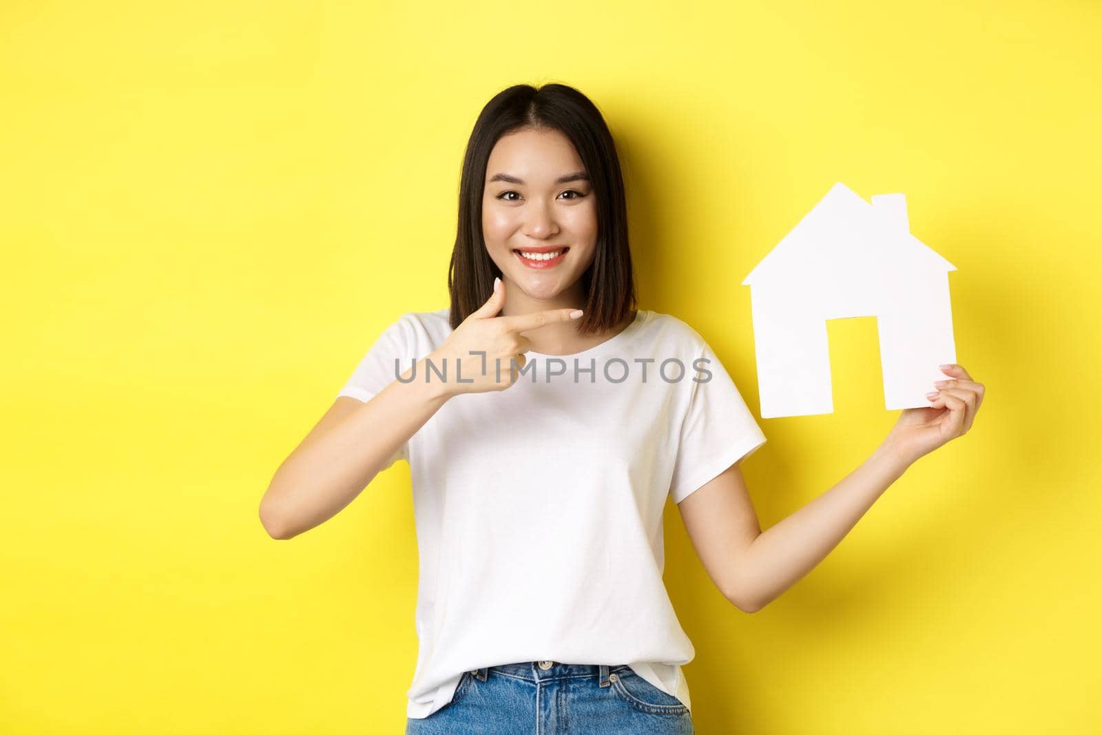 Real estate and insurance concept. Cheerful asian woman smiling, pointing at paper house cutout, recommend agency logo, standing over yellow background by Benzoix
