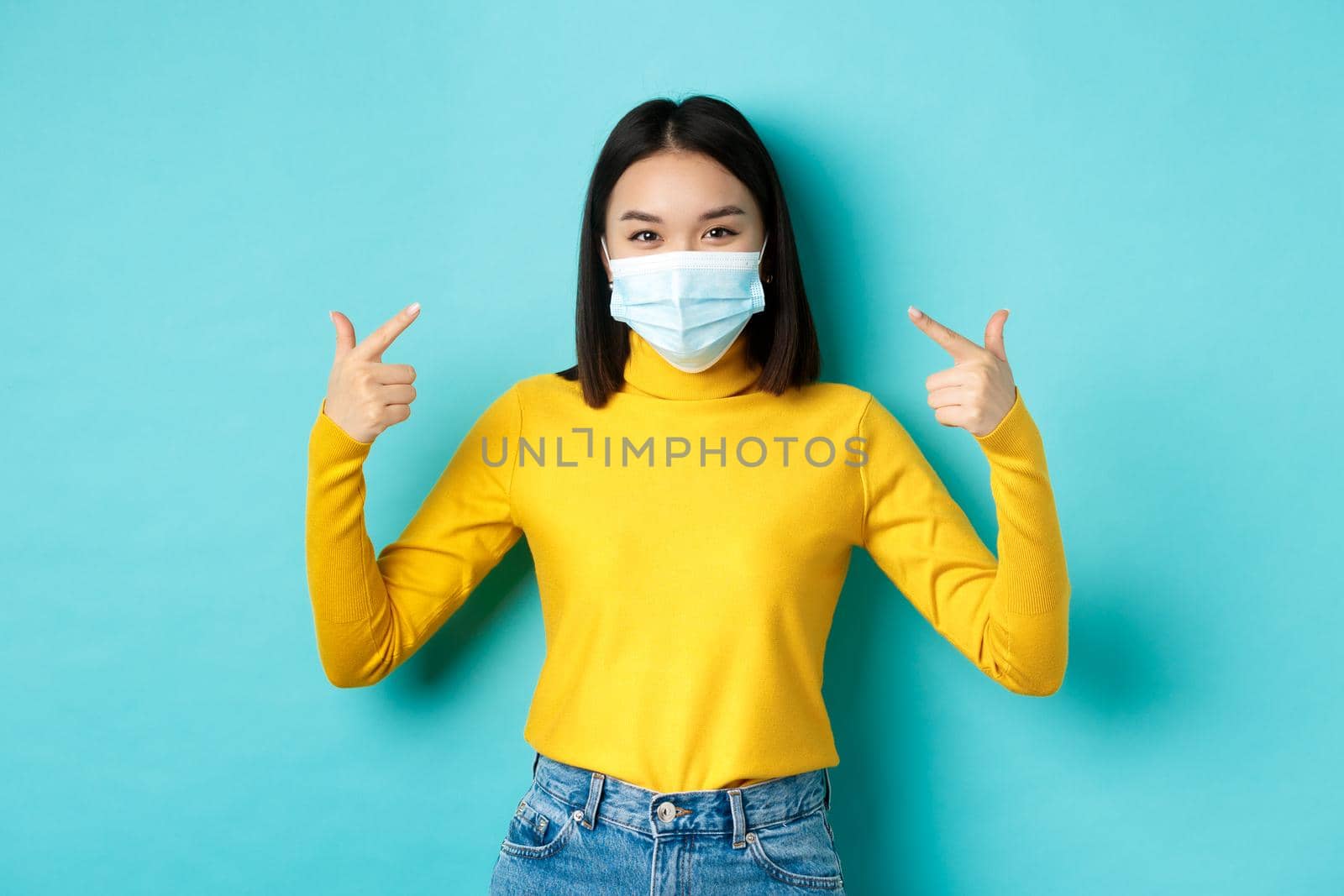 Covid-19, social distancing and pandemic concept. Young asian woman protect herself from coronavirus, pointing finger at her medical mask, standing over blue background.