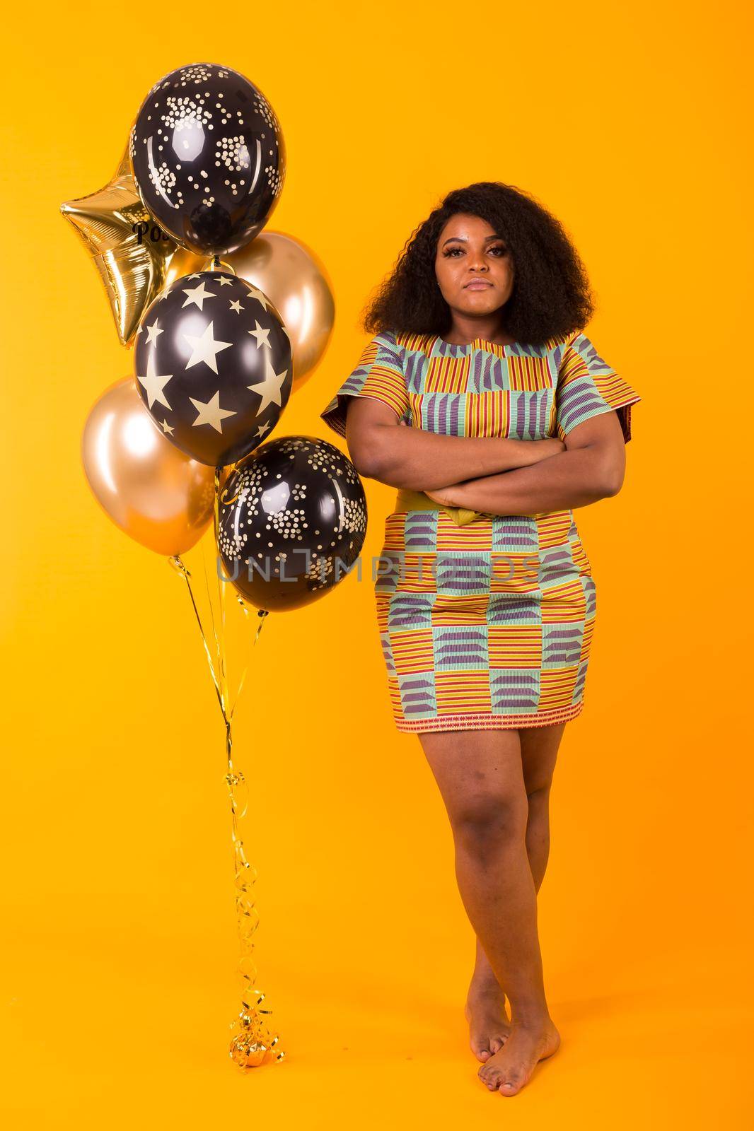 Portrait of smiling young African-American adult woman looking sweet on yellow background holding balloons.