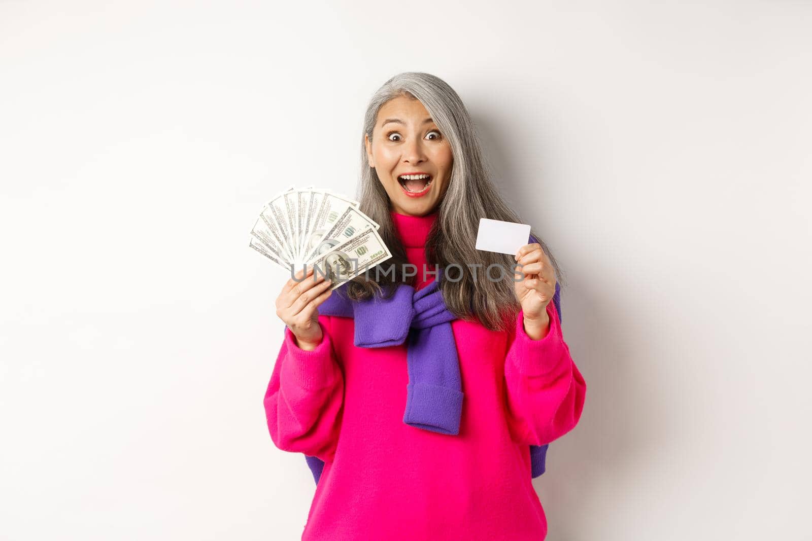 Shopping concept. Amazed asian granny showing plastic credit card and money dollars, want to buy something, standing over white background by Benzoix