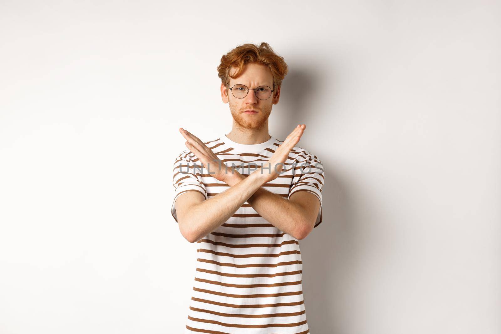 Serious young man with ginger hair frowning, showing stop gesture, making cross to prohibit something bad, disagree with you, standing over white background by Benzoix