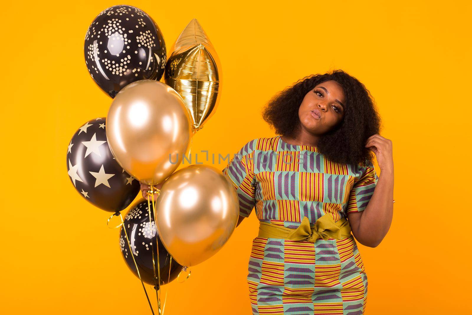 Portrait of smiling young African-American adult woman looking sweet on yellow background holding balloons.