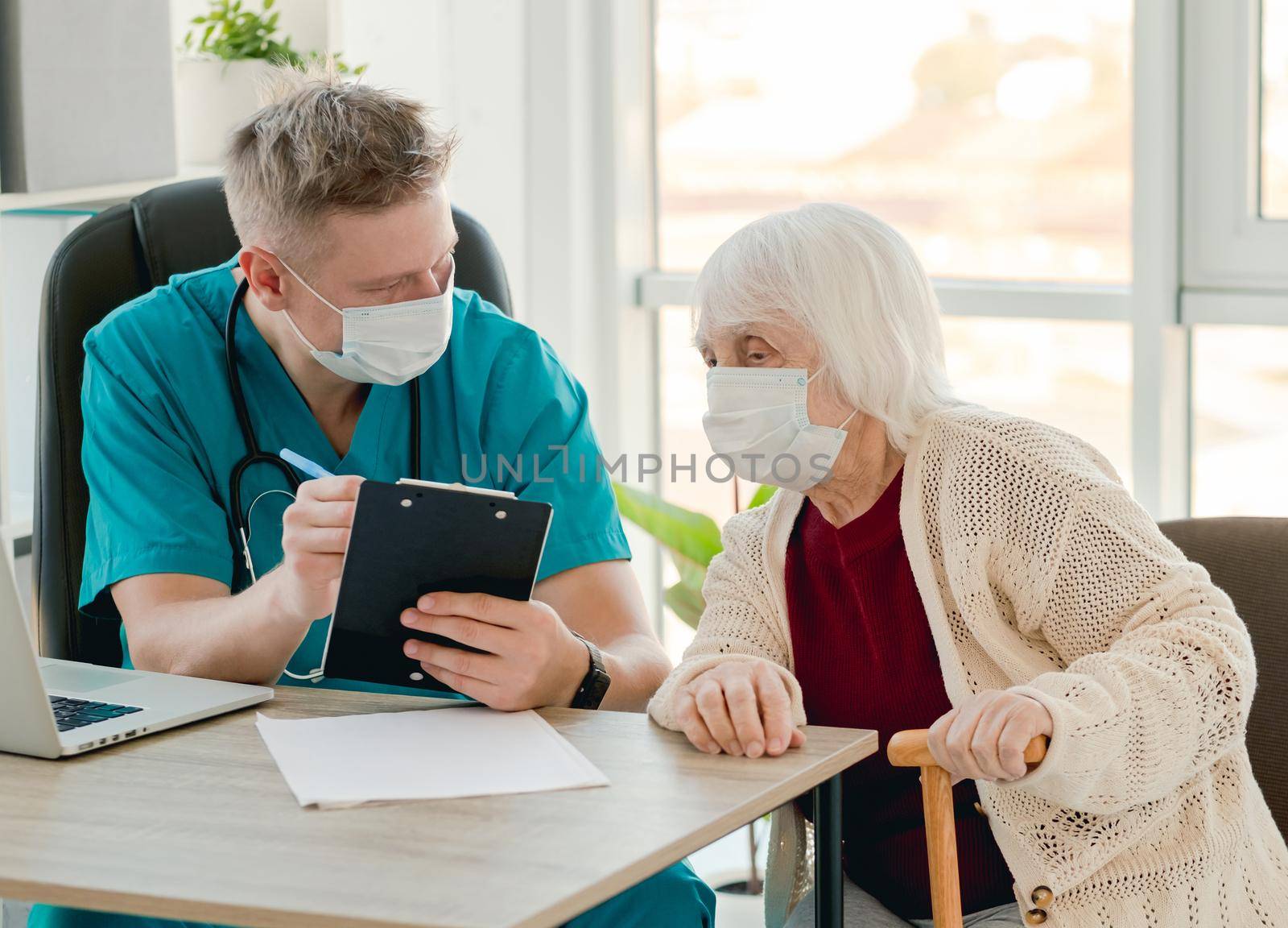 Doctor in protective face mask writing elderly patient complaints during appointment in clinic