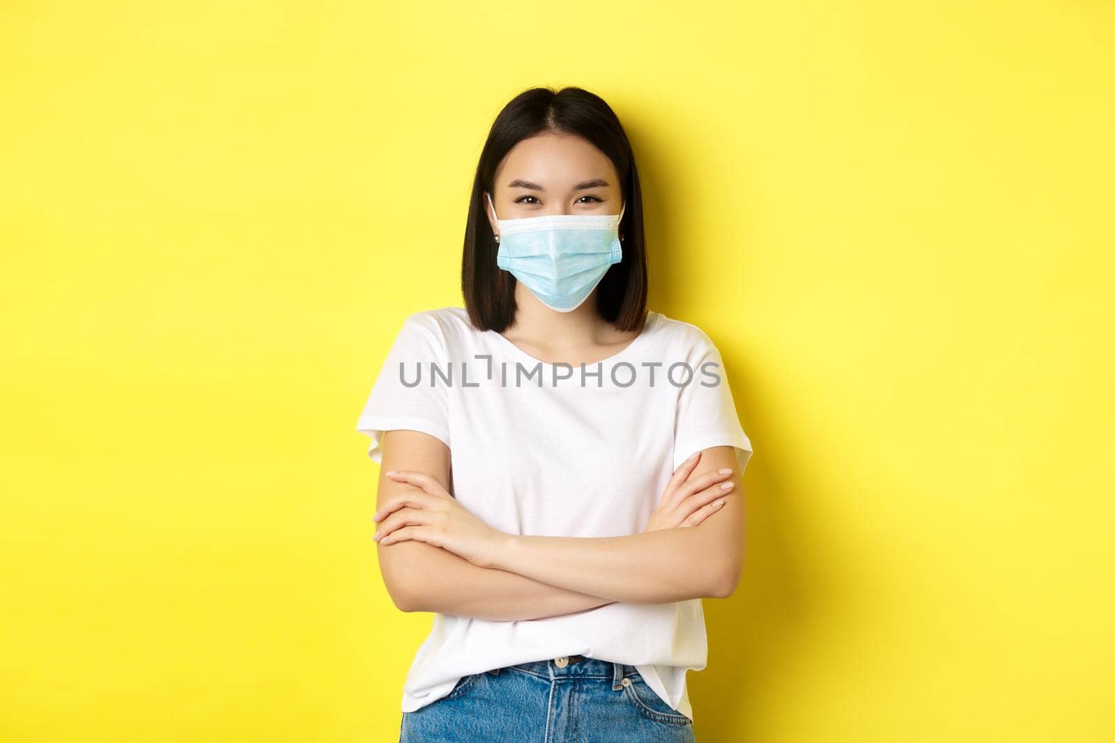Covid, health care and pandemic concept. Asian woman in white t-shirt and medical mask cross arms on chest and looking at camera.
