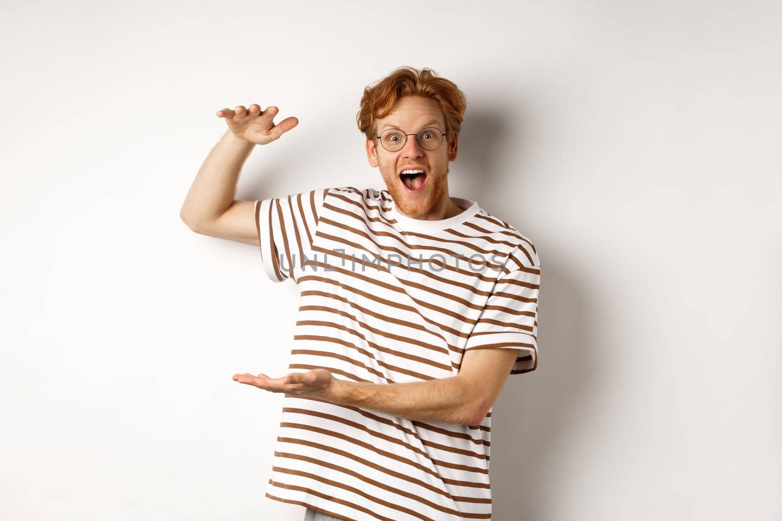 Amazed redhead man in glasses showing something large size and smiling at camera, saying wow, standing over white background.