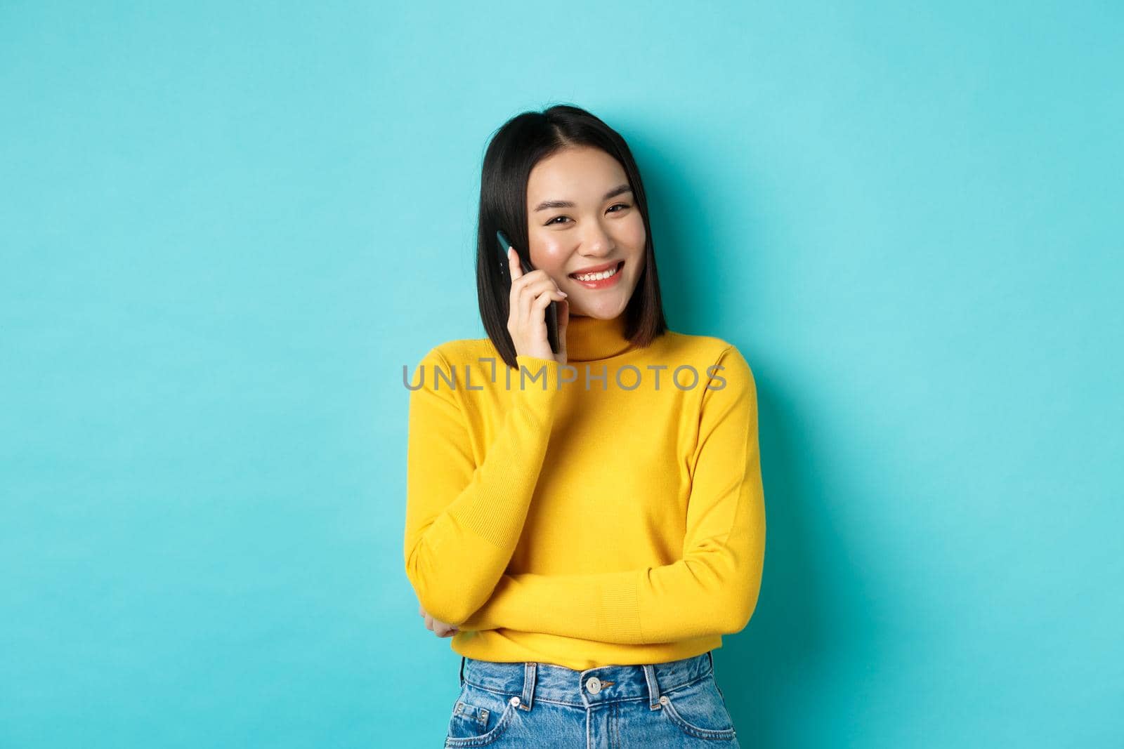 Young stylish asian woman talking on phone, calling friend and smiling, standing with smartphone against blue background by Benzoix