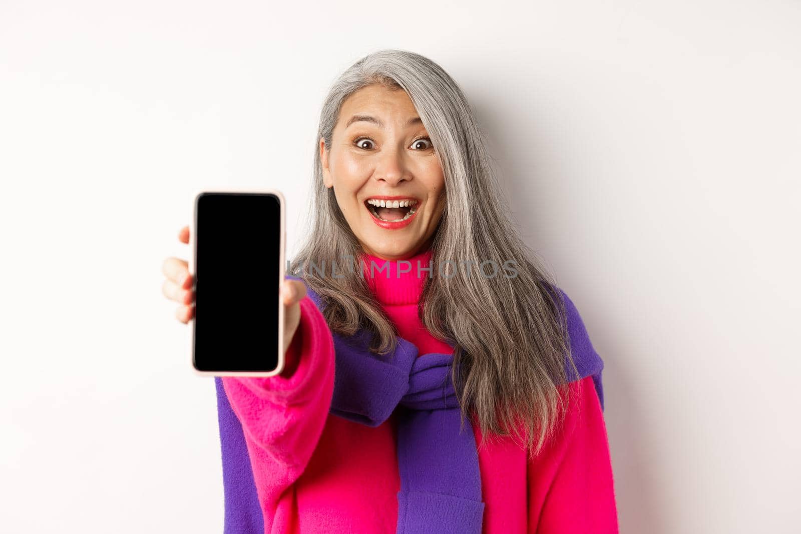 Online shopping. Close up of stylish asian senior woman extending hand with mobile phone, showing blank smartphone screen and smiling, standing over white background by Benzoix