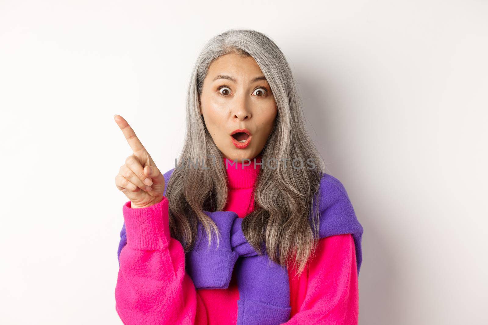 Fashion. Close up of amazed asian senior woman pointing finger upper left corner, gasping and looking impressed at camera, standing over white background.
