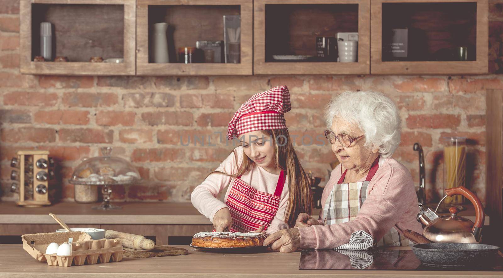 Granddaughter with her grandmother sitting at the kitchen and cut the homemade pie