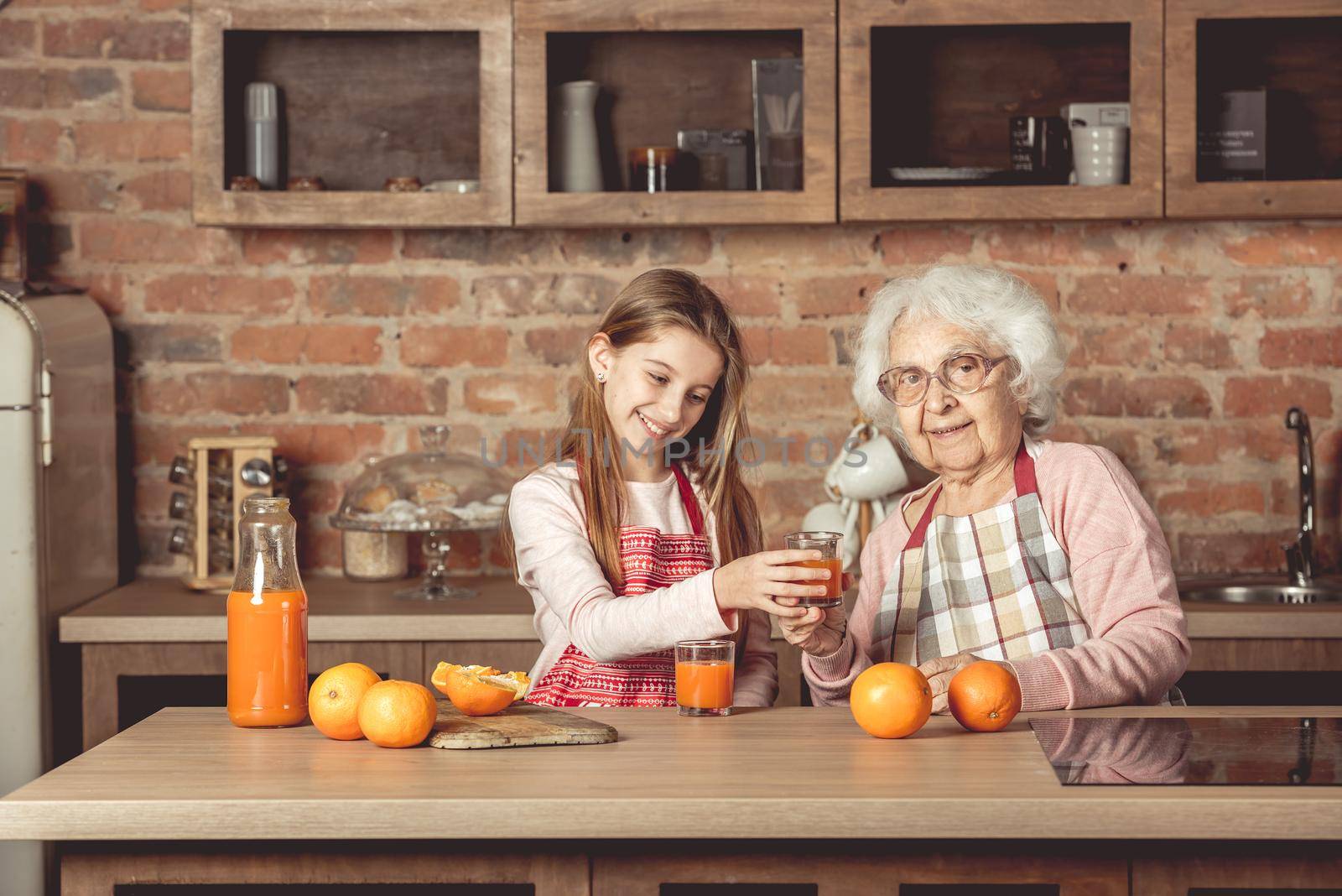 Granny with happy granddaughter tasting and drinking fresh natural orange juice at kitchen at home