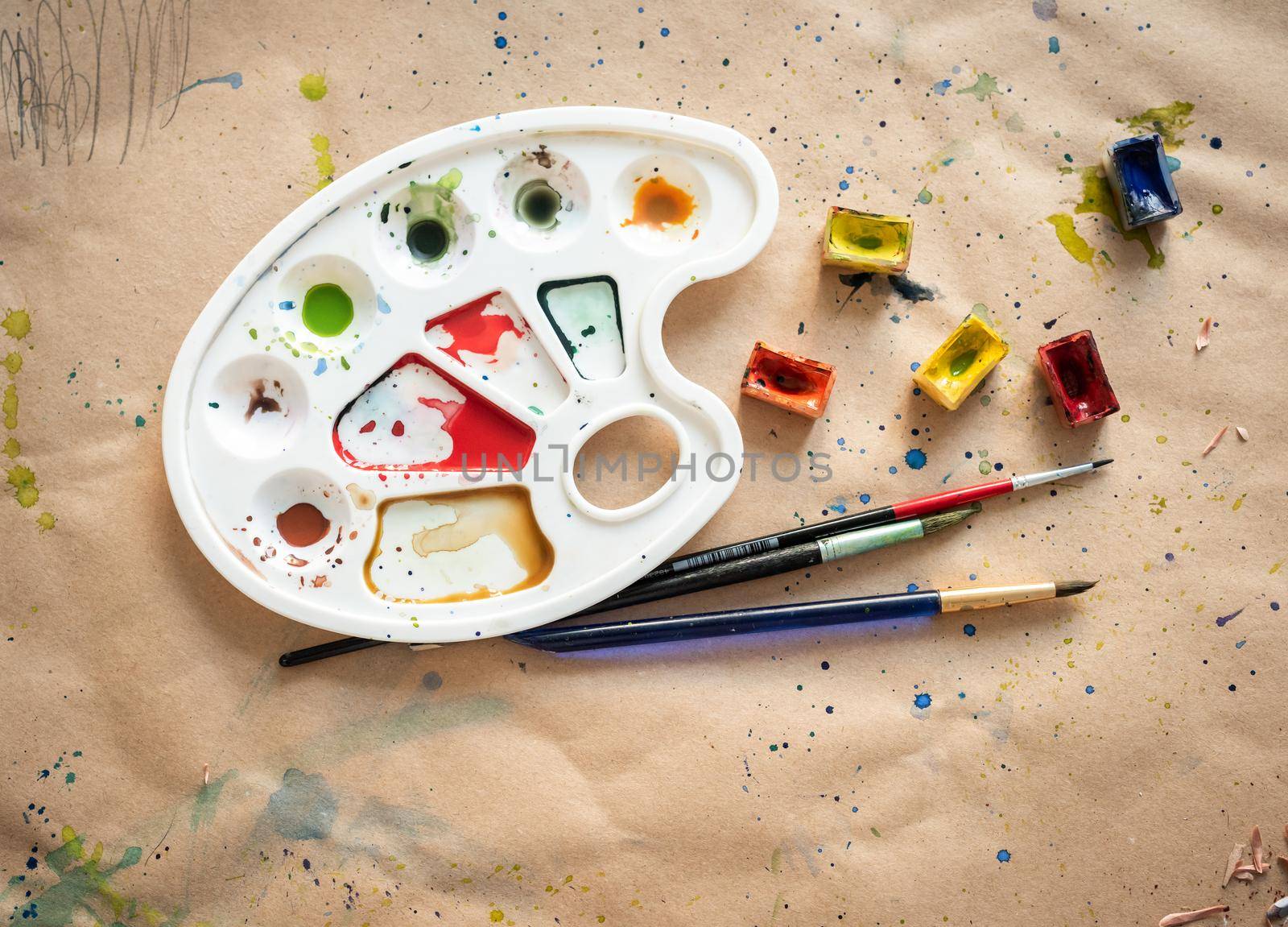 Plastic art palette with used brushes and opened paint boxes on paper-covered workshop table