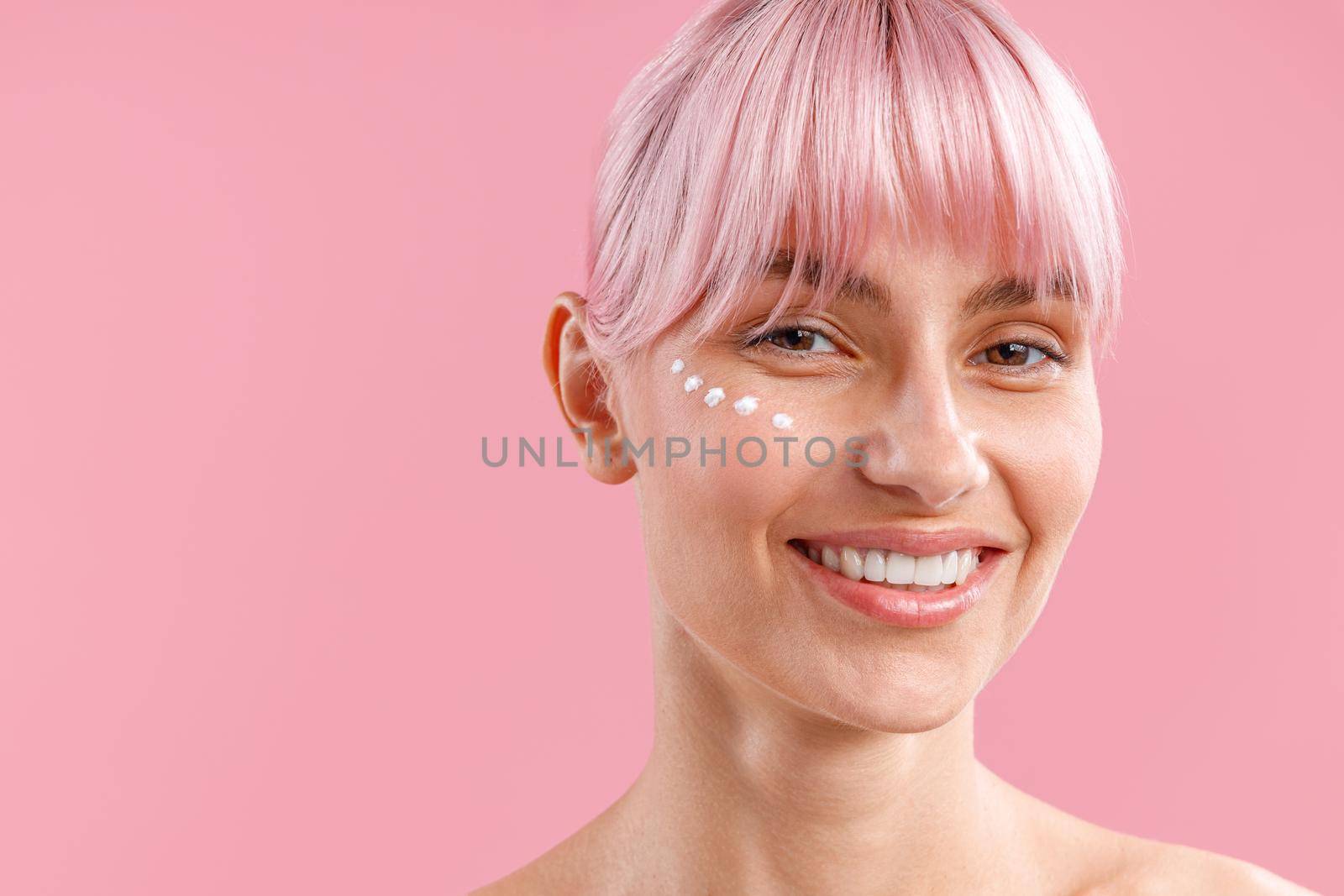 Portrait of smiling woman with pink hair and nourishing cream applied on her face like dots isolated over pink background. Beauty, spa, skin care concept