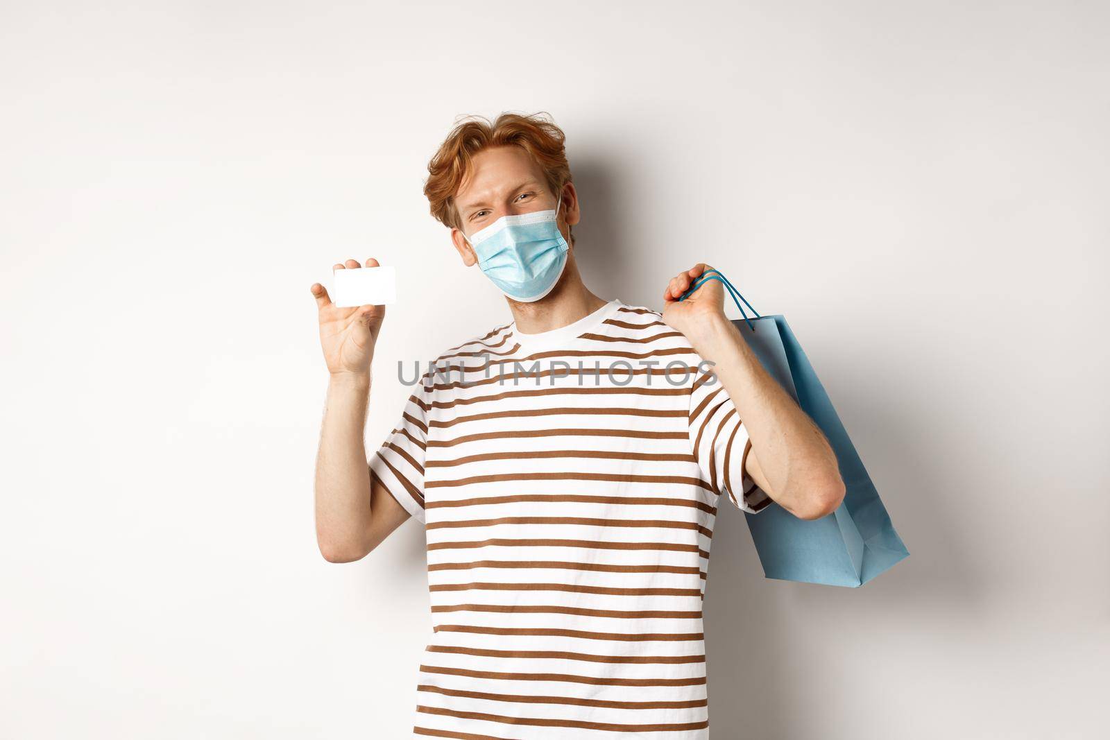 Concept of covid-19 and lifestyle. Cheerful young man with red hair, wear medical mask, showing shopping bag from store and plastic credit card by Benzoix