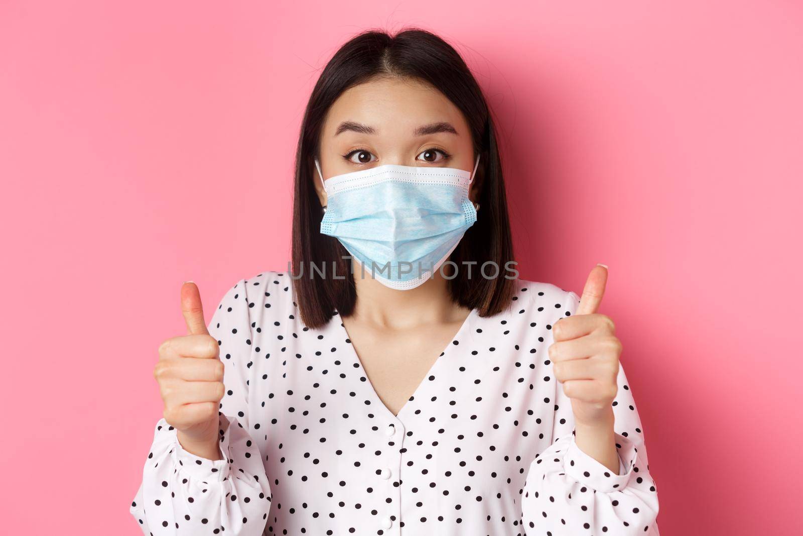 Covid-19, pandemic and lifestyle concept. Supportive asian female in face mask showing thumbs-up, approve and praise good work, standing over pink background by Benzoix
