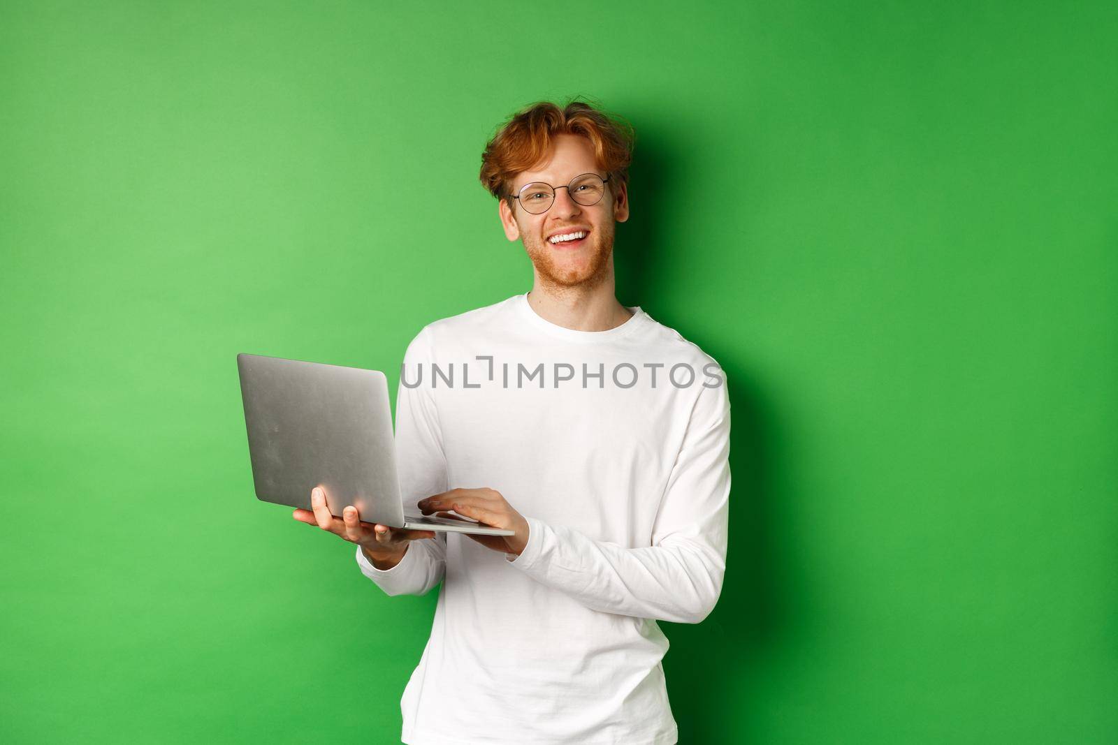 Cheerful redhead freelancer in glasses smiling at camera, working on laptop while standing over green background.