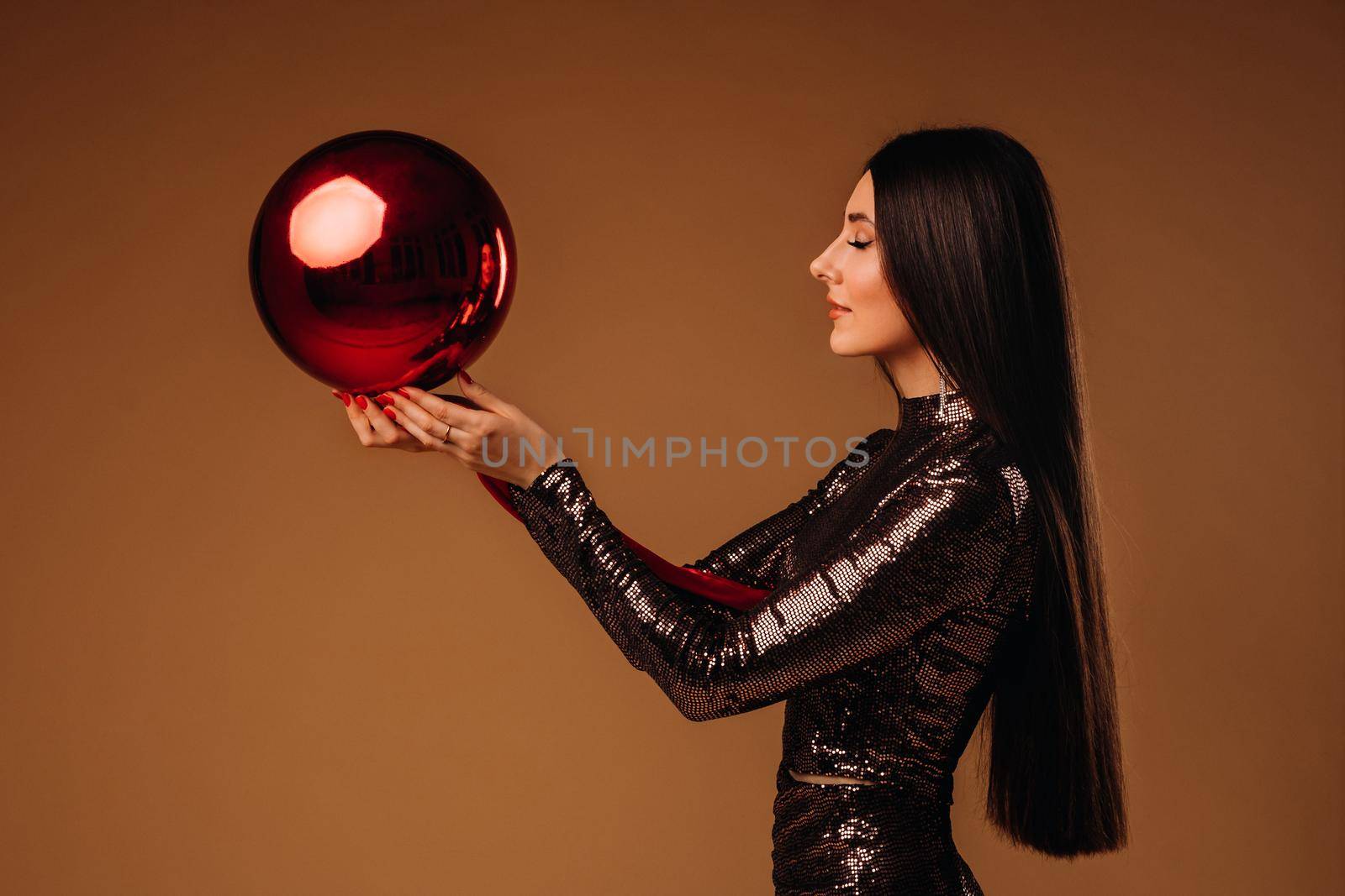 Girl with long hair in a shiny dress with a large Christmas ball on a brown background.