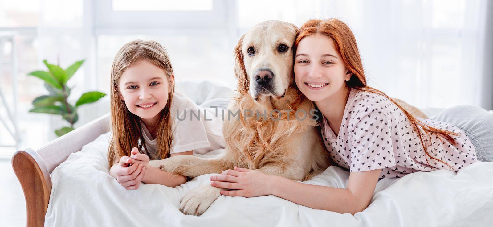 Two sisters lying in the bed with golden retriever dog in the morning time and looking at the camera. Girls with pet staying at home. Beautiful portrait of friendship between human and animal