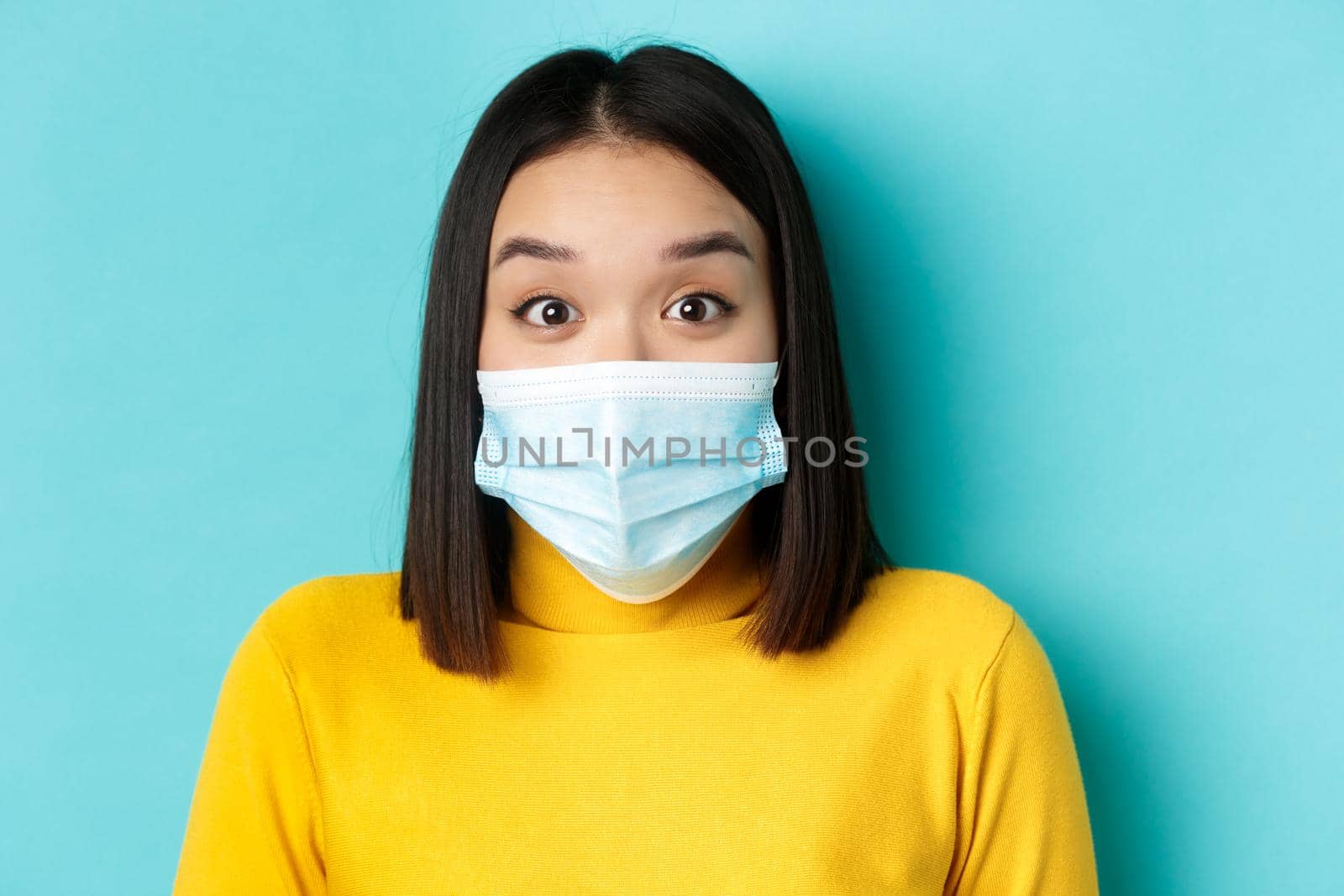 Covid-19, social distancing and pandemic concept. Close up of surprised asian woman in medical mask, raise eyebrows and look amazed at camera, blue background.