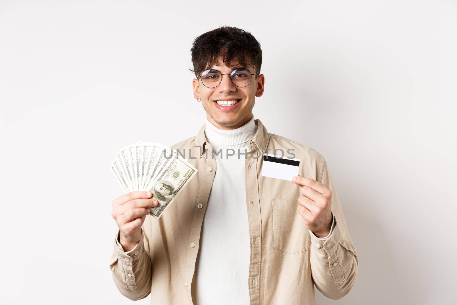 Happy person stands with money and plastic credit card. Young man showing cash and smiling satisfied, standing on white background.