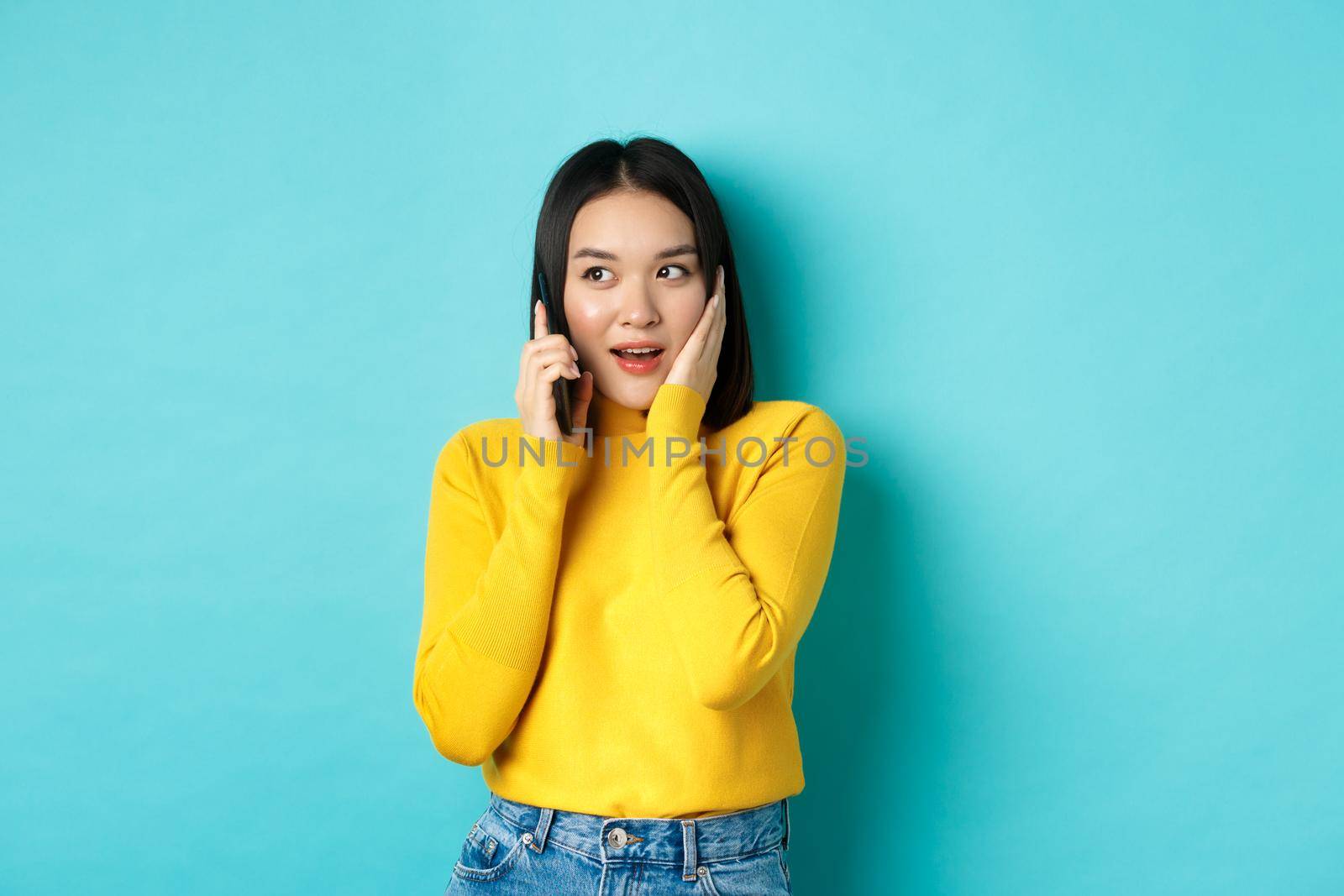 Woman looking intrigued while talking on mobile phone, receive interesting offer during call, standing over blue background.