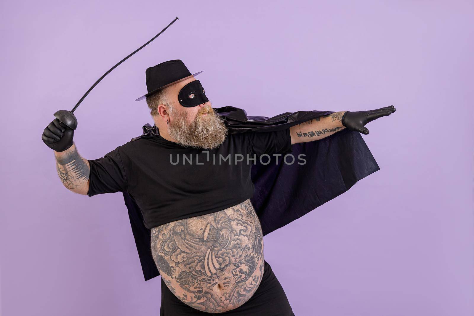Mature bearded fat man in Zorro costume with large bare abdomen stands in bellicose pose on purple background in studio