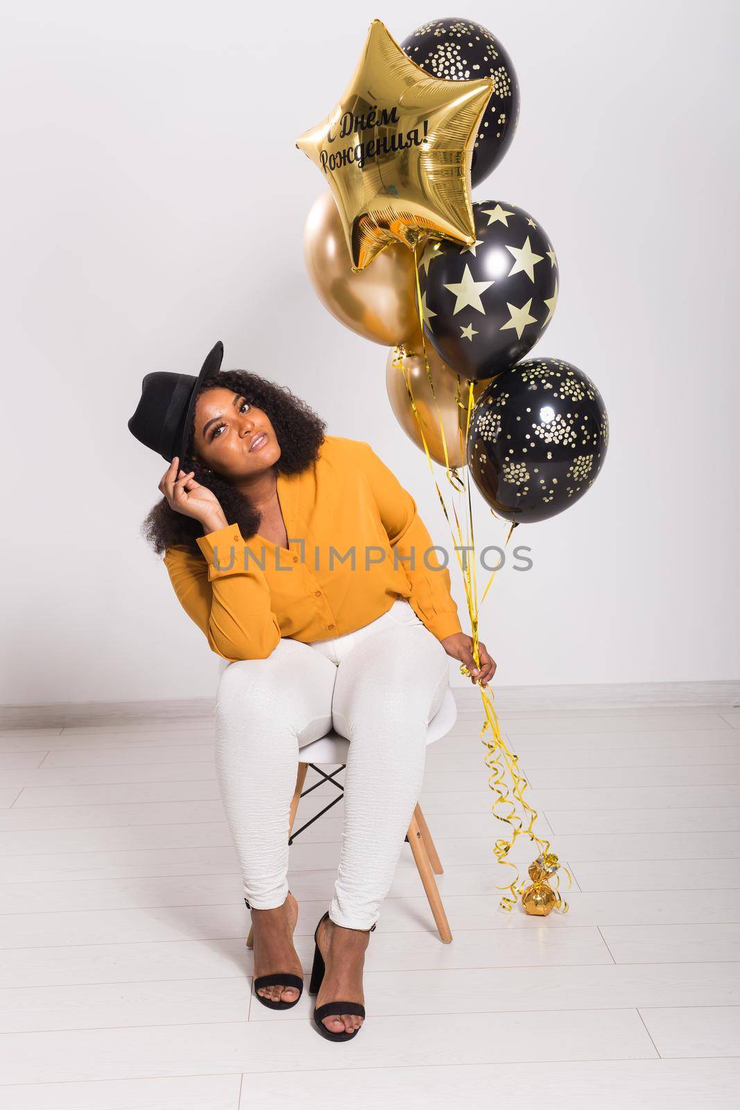 Portrait of smiling young African-American adult woman looking sweet on yellow background holding balloons.