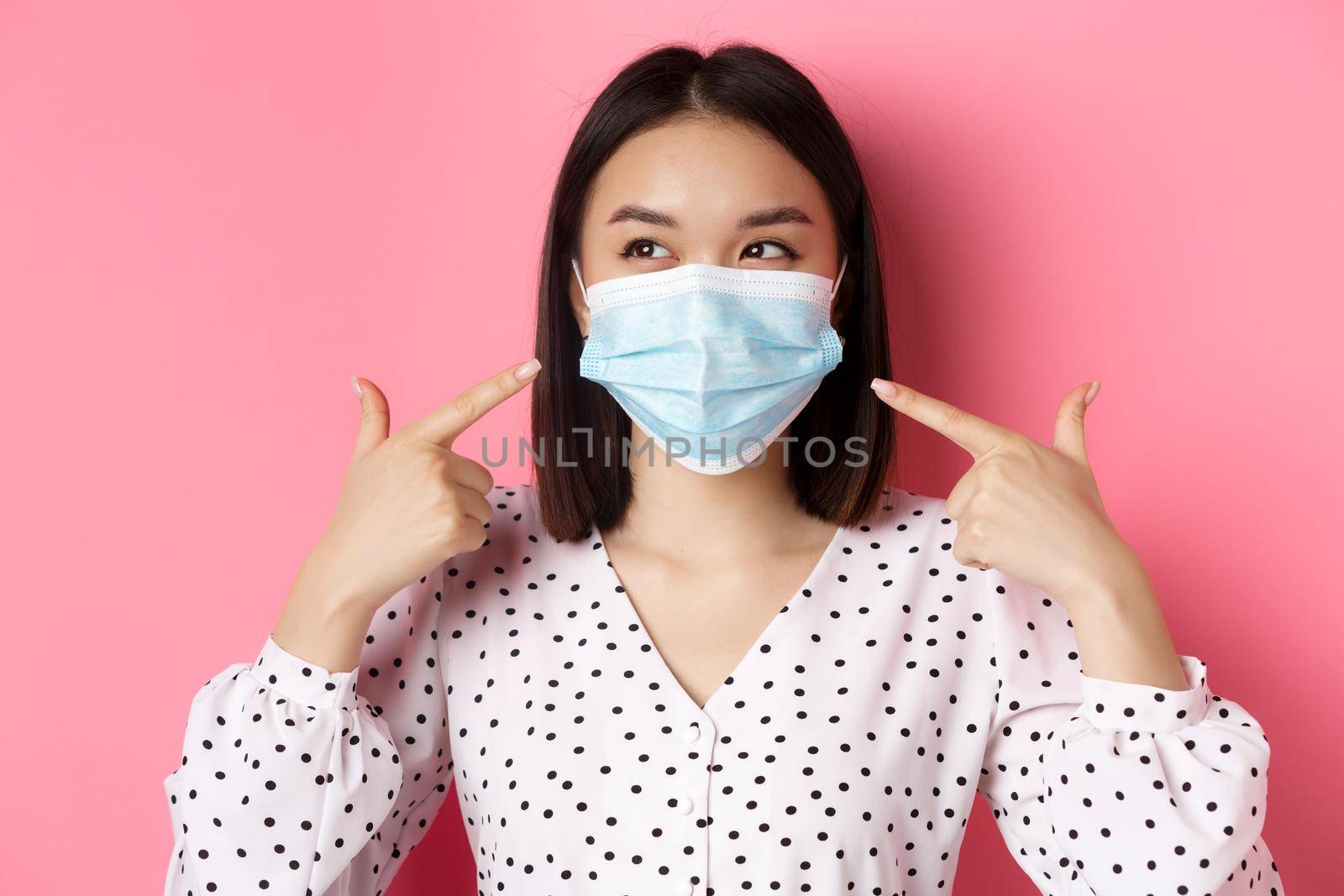 Covid-19, pandemic and lifestyle concept. Kawaii asian girl pointing fingers at her face mask, wearing preventive measures from coronavirus, smiling with eyes, pink background.