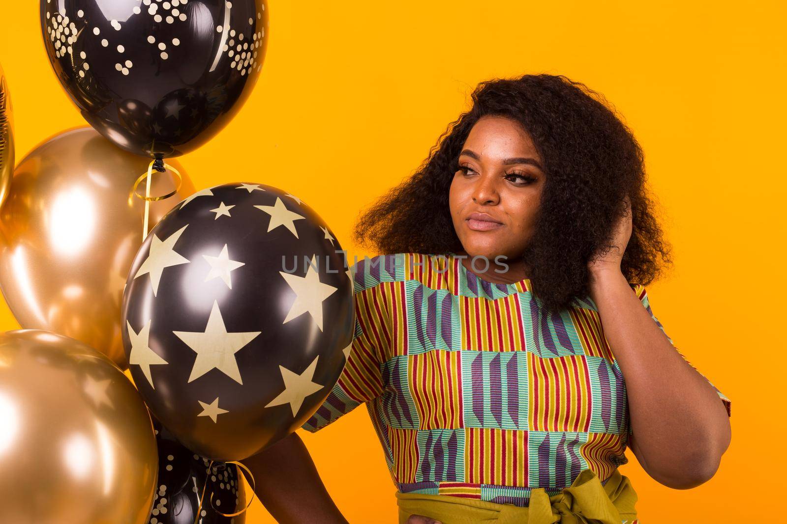 Portrait of smiling young African-American adult woman looking sweet on yellow background holding balloons.