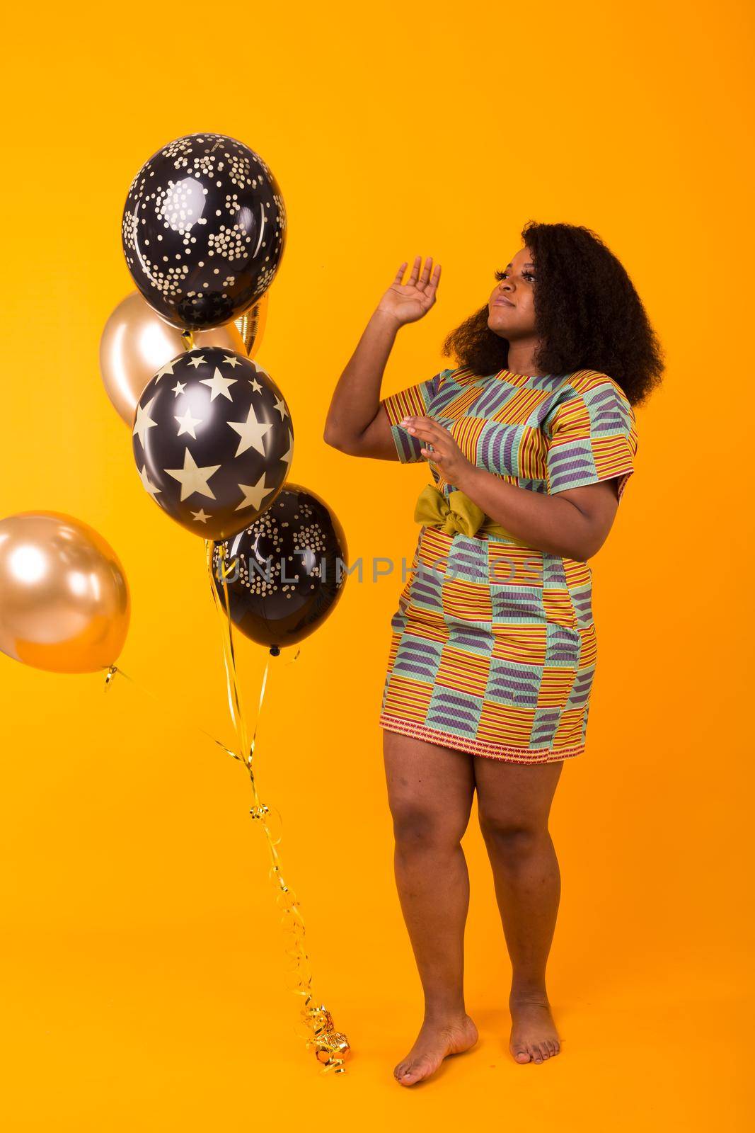 Holidays, party and fun concept - Portrait of smiling young African-American young woman looking sweet on yellow background holding balloons. by Satura86