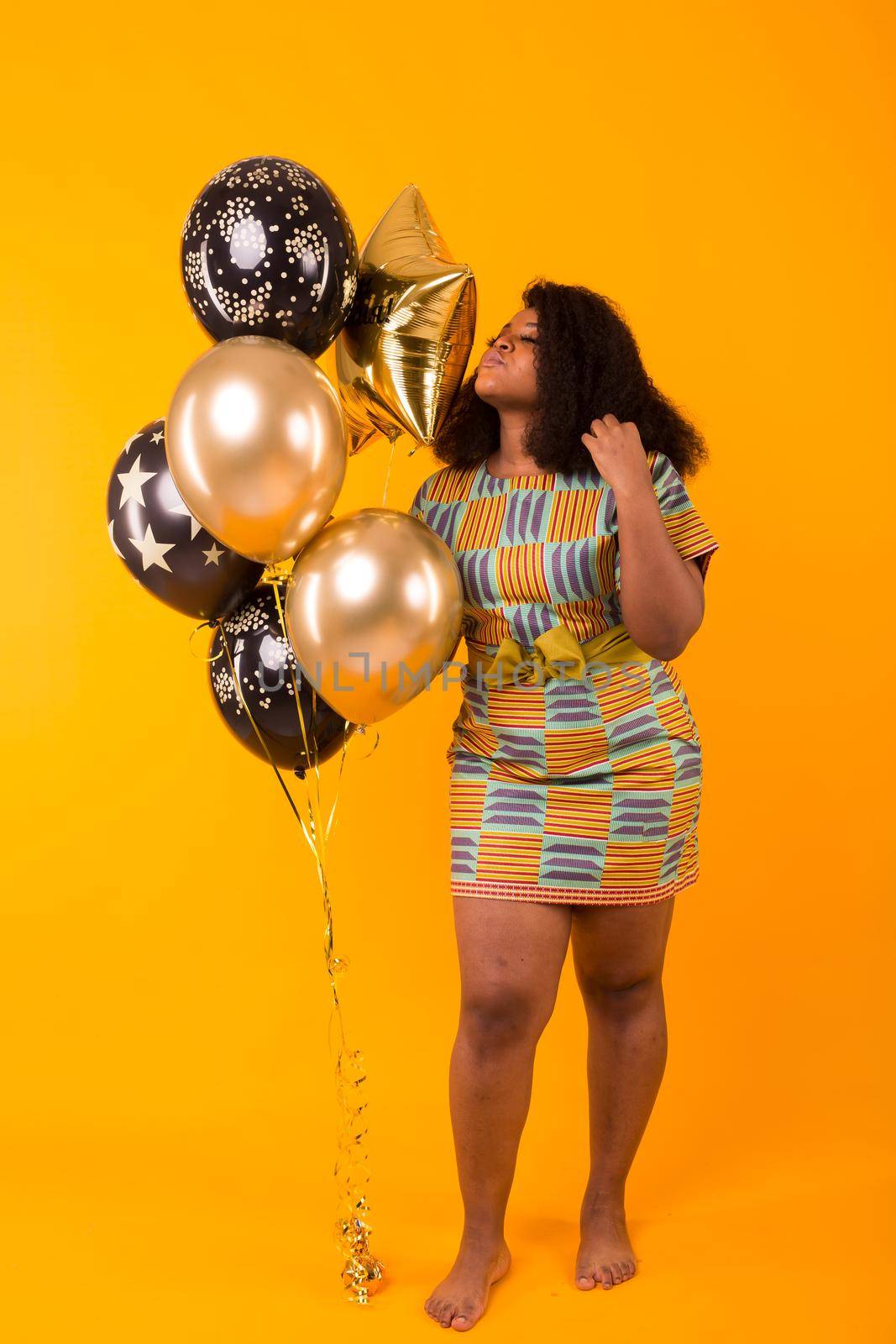 Holidays, birthday party and fun concept - Portrait of smiling young African-American young woman looking sweet on yellow background holding balloons. by Satura86