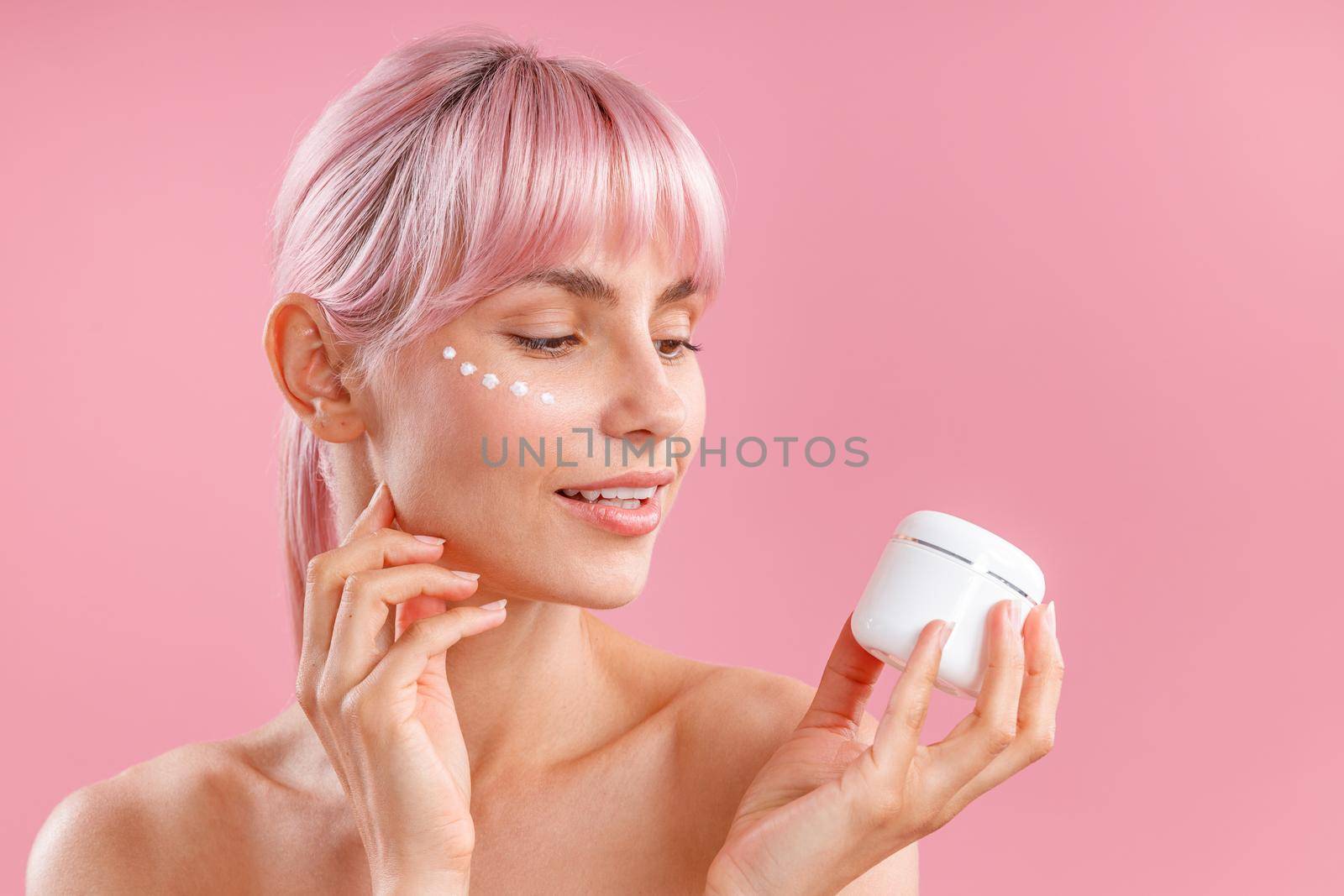 Portrait of beautiful woman with pink hair and face cream applied on her skin like dots holding and looking at cream in white jar isolated over pink background by Yaroslav_astakhov