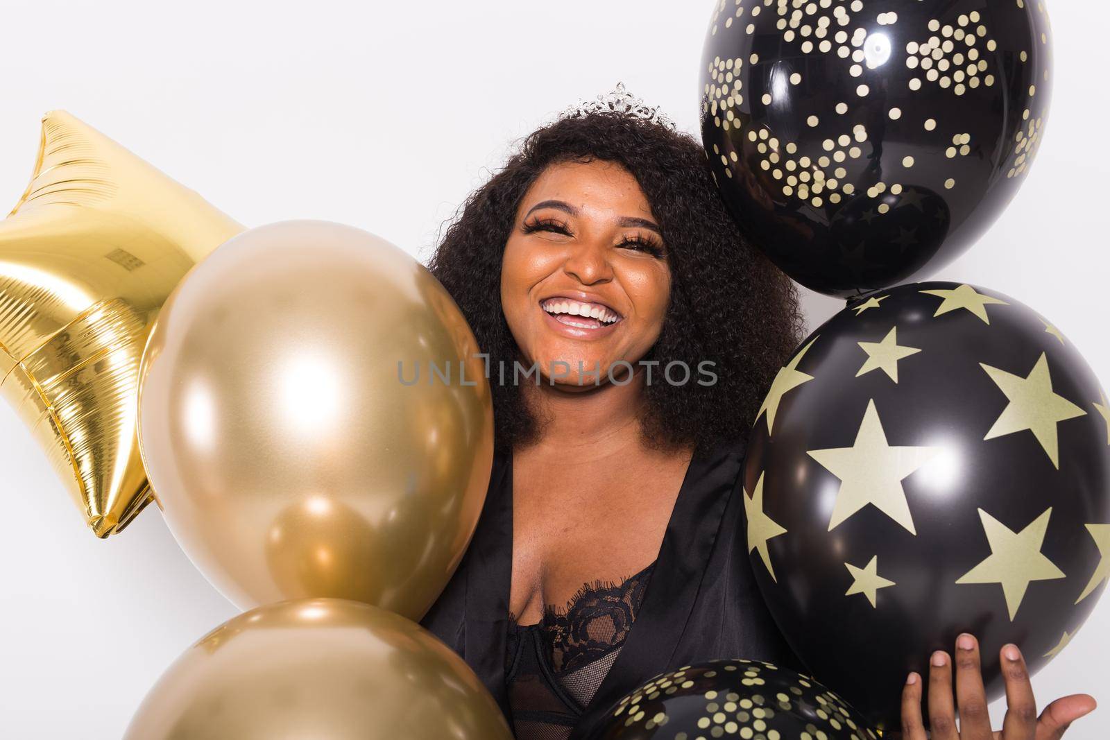 Portrait of smiling young African-American adult woman looking sweet on yellow background holding balloons.