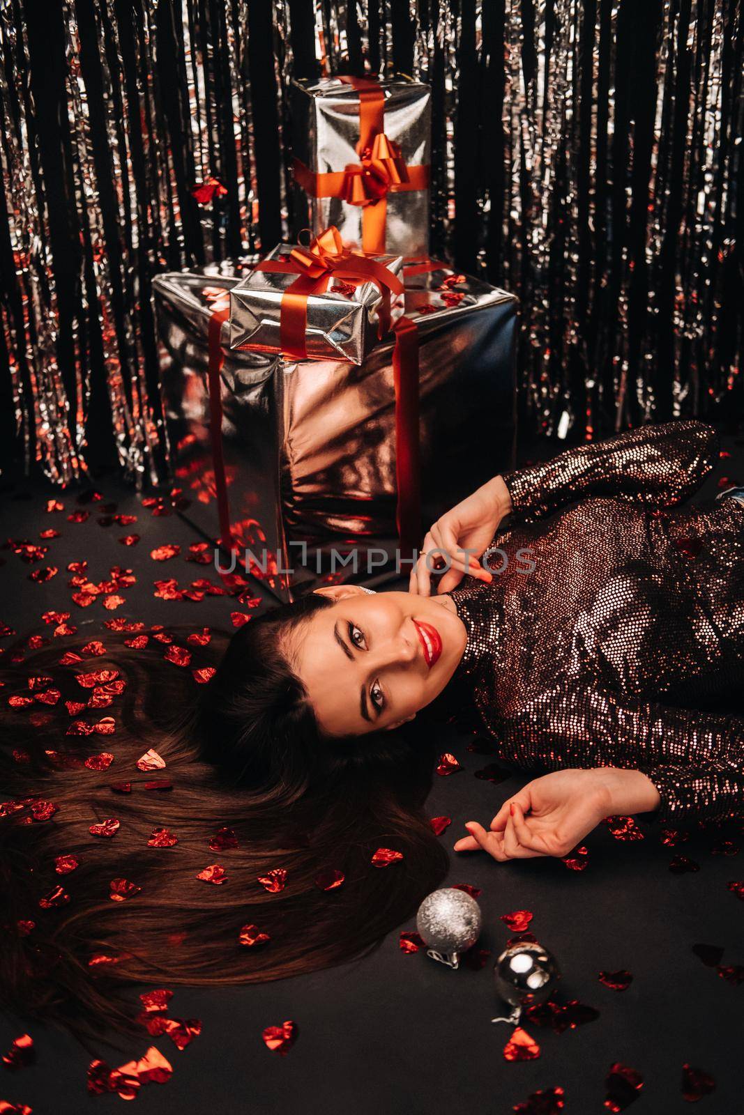 Top view of a girl lying in shiny clothes on the floor in confetti in the form of hearts and gifts.