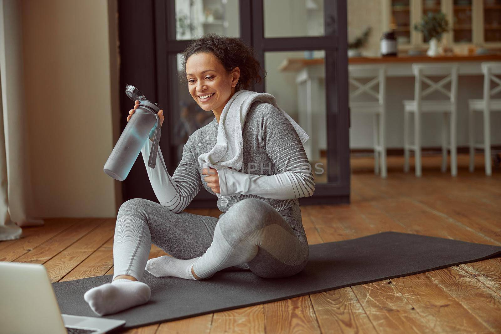Merry slim woman with towel is drinking water after workout while sitting on mat before laptop